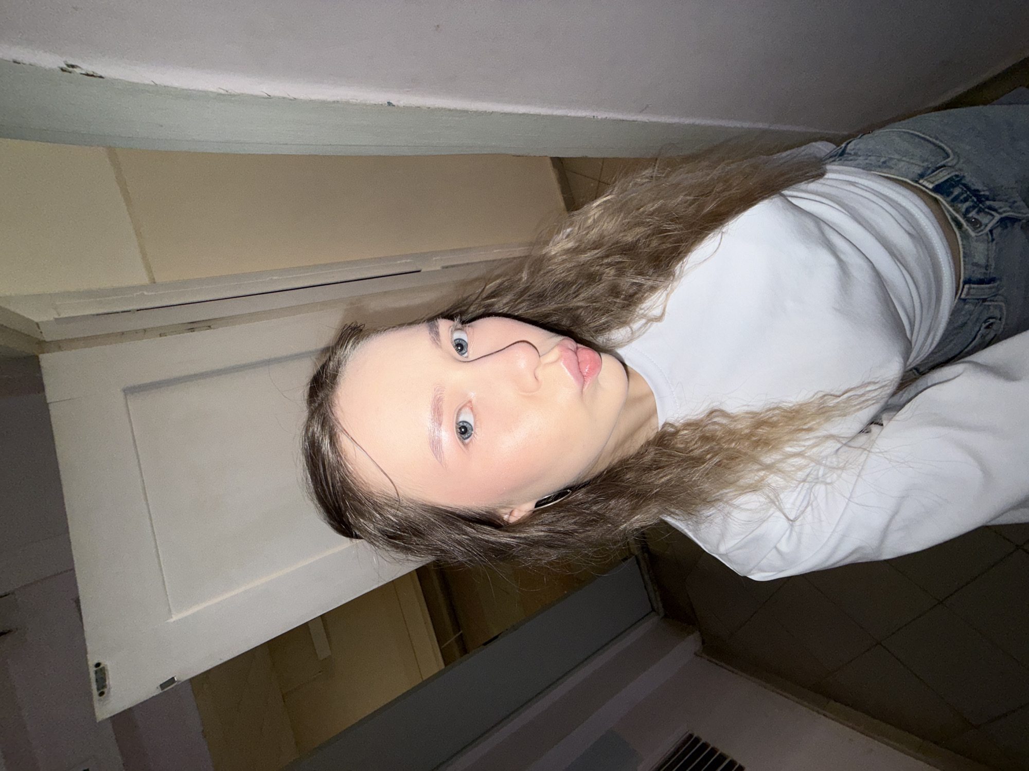 The media file displays a young woman posing indoors, with the image rotated 90 degrees counter-clockwise, making her appear sideways in the frame. She has long, wavy light brown or blonde hair, blue eyes, and light makeup featuring pink blush. Her lips form a slight pout as she looks towards the camera. She is wearing a white long-sleeved top and light-wash denim jeans. A small, dark earring or ear cuff is visible on her right ear.
The setting is a simple indoor space, possibly a hallway or room. The background includes walls painted in varying light shades, such as cream or light yellow and a lighter, potentially light blue or grey, section. A white door or cabinet door with a plain panel design is partially visible on the left, showing some signs of wear, including a visible latch mechanism. The ceiling edge above the door frame also exhibits some minor imperfections. The floor is covered with dark square tiles, and a vent or grille is present in the lower right portion of the image. The lighting appears to be artificial and somewhat dim. No visible text is present. The overall scene suggests a casual indoor moment in Chaikovsky, Russia.