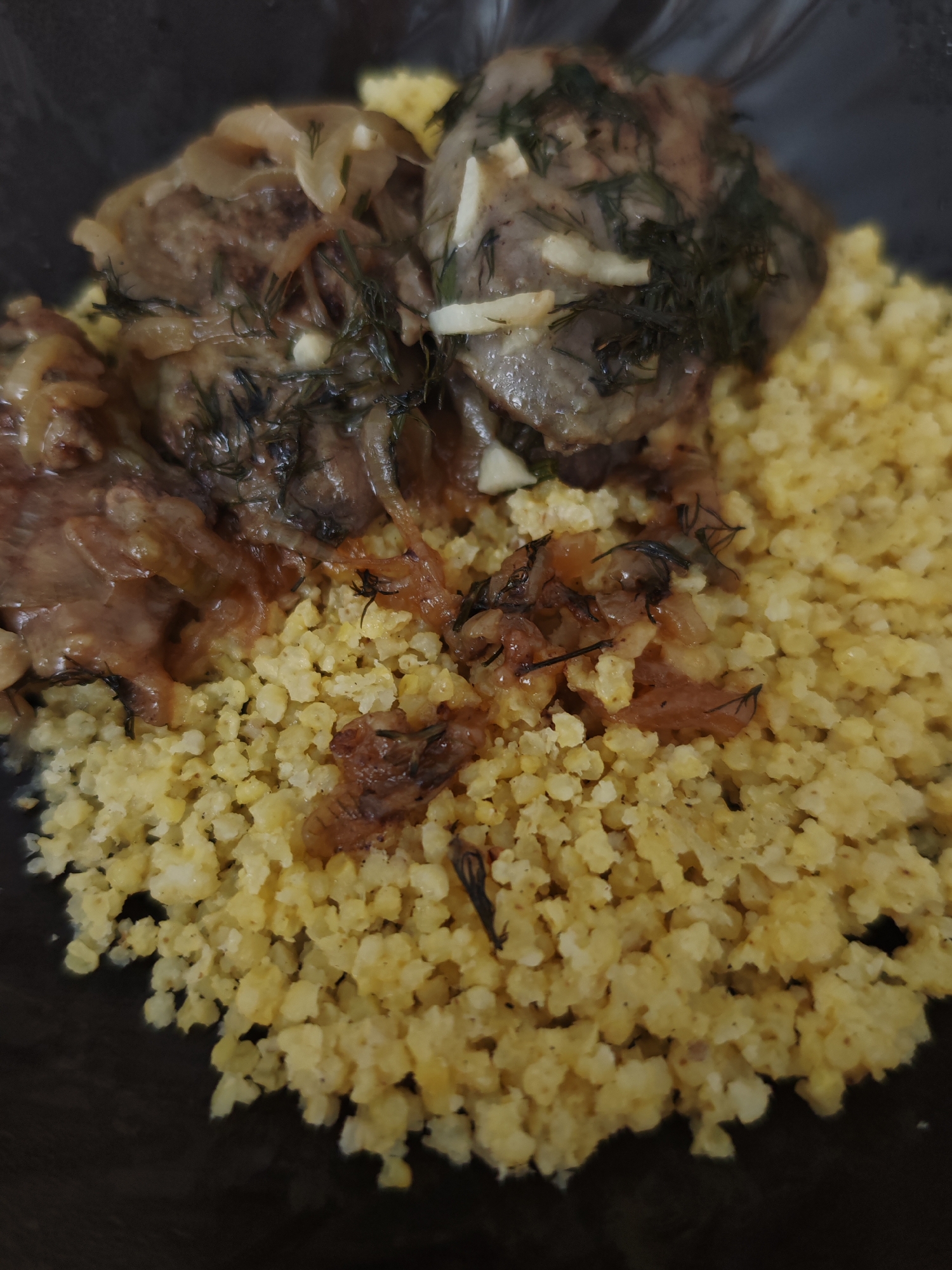 A close-up shot shows a serving of food in a dark bowl. The dish consists of a bed of light yellow, granular food, possibly couscous or polenta, topped with pieces of meat. The meat appears to be braised or stewed, with visible slices of onion and sprigs of fresh dill. Some of the meat pieces are covered in a savory sauce. The overall presentation suggests a home-cooked meal. The lighting is somewhat dim, hinting at indoor dining. There is no visible text in the image.