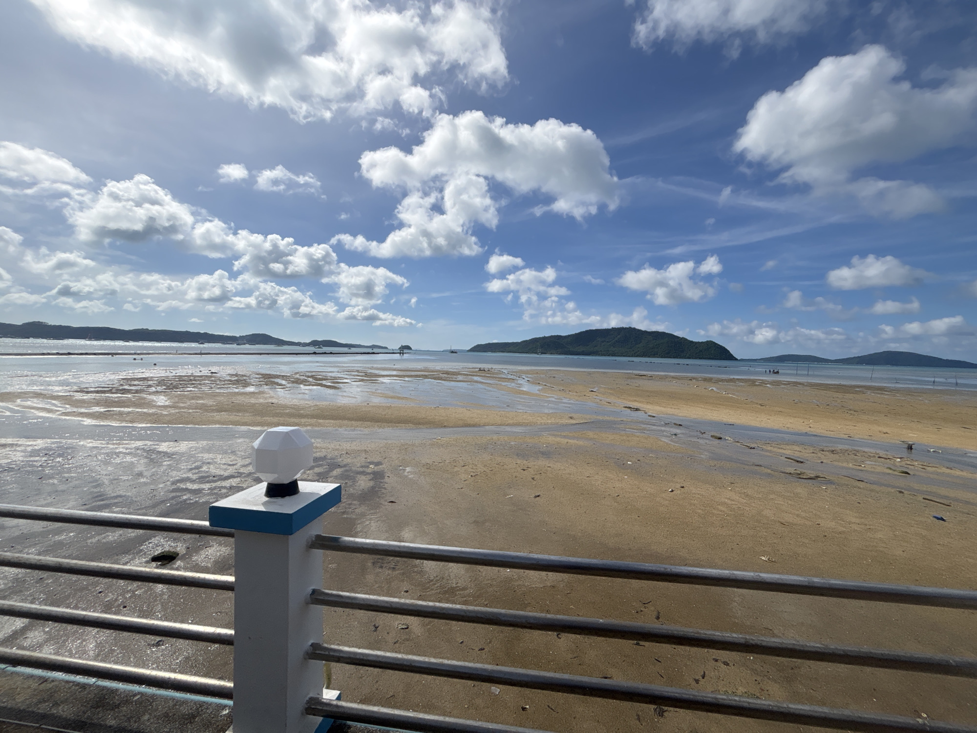 A wide-angle, eye-level view captures a sandy beach at low tide under a bright, partly cloudy blue sky. In the foreground, a metal railing with horizontal bars runs across the bottom of the frame. To the left of the railing, a white pillar topped with a geometric lantern stands. The pillar has blue trim along its top edge. Beyond the railing, the expansive beach is a mix of wet sand, reflecting shallow water, and dry sand, with streaks of dark seaweed or debris scattered across it. In the distance, several small islands with green, tree-covered hills rise from the water. A few boats are visible on the horizon, and a small group of people can be seen walking on the beach in the far distance. The scene appears to be at Ao Sane Beach in Rawai, Thailand, judging by the characteristic landscape of islands and the appearance of the beach during low tide. The lighting suggests it is daytime, possibly late morning or early afternoon, with the sun casting some shadows but overall illuminating the scene brightly.