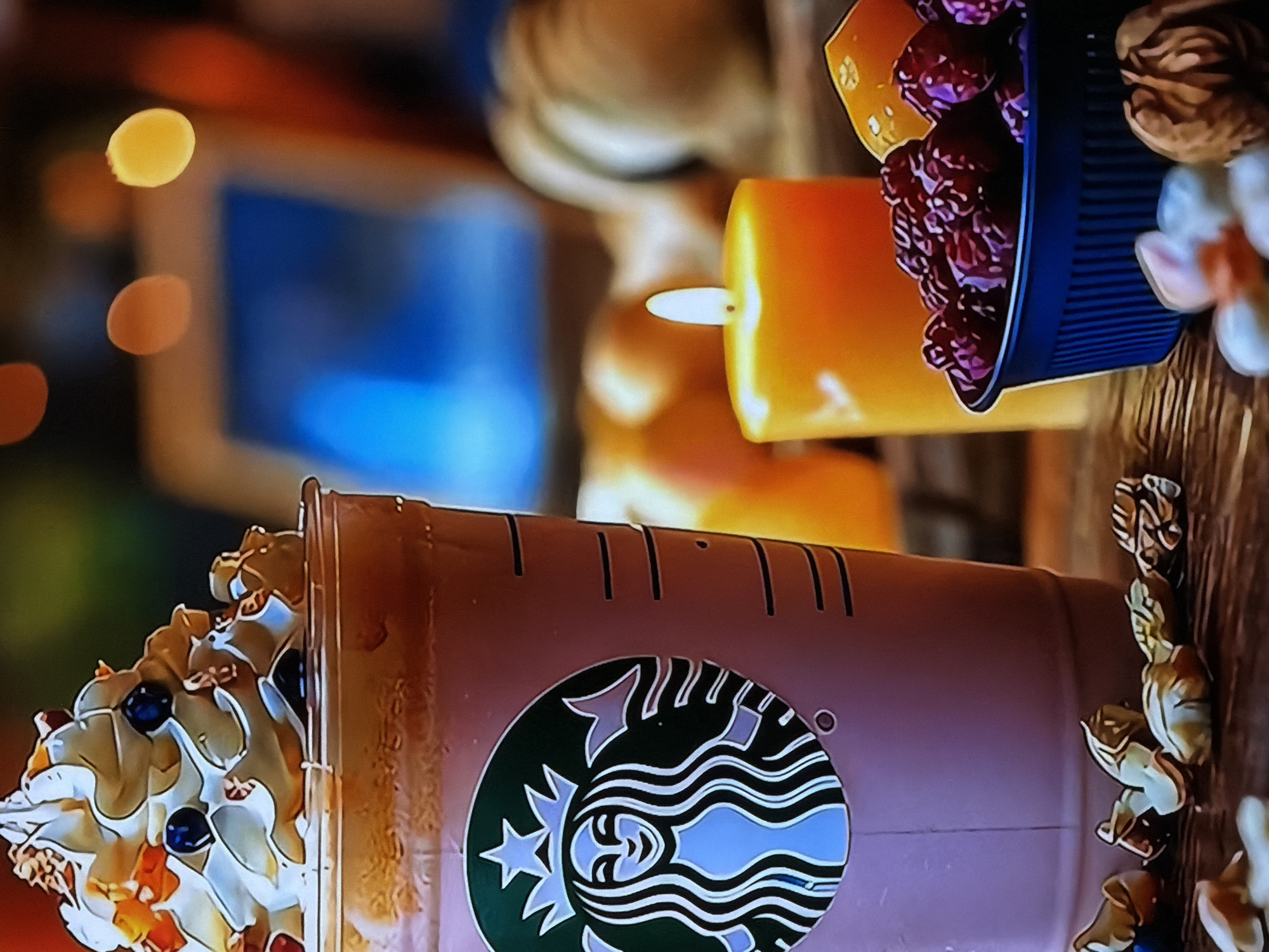 The image displays a close-up, diagonally oriented view of a Starbucks-branded drink cup, positioned towards the lower left of the frame. The cup is a muted, light brown or pinkish color, prominently featuring the iconic Starbucks Siren logo in green, white, and light blue. The beverage inside is topped with a generous swirl of whipped cream, garnished with dark blueberries and smaller, light-colored pieces, possibly nuts or dried fruit.
Adjacent to and above the cup, a warm orange cylindrical candle casts a soft glow with its visible flame. To the right of the candle, a small, dark blue ridged container holds a collection of deep red dried fruits, likely cranberries. Scattered around the candle and container on a wooden surface are several whole walnuts, contributing to a rustic aesthetic.
The background is softly blurred with bokeh effects, showing out-of-focus warm yellow and orange lights. A distinct, blurred rectangular blue shape, possibly a screen or window, is also visible. The overall setting appears to be indoors, suggesting a cozy or festive atmosphere, perhaps in a cafe or home environment. The arrangement of the items, including the lit candle, berries, and walnuts, evokes a warm, comforting, and possibly seasonal (autumnal or winter) mood. No people are visible, and no specific activity is taking place; it is a still-life composition. The primary visible text is the Starbucks logo. The location context for this media file is Sofiivska Borshchahivka, Ukraine.