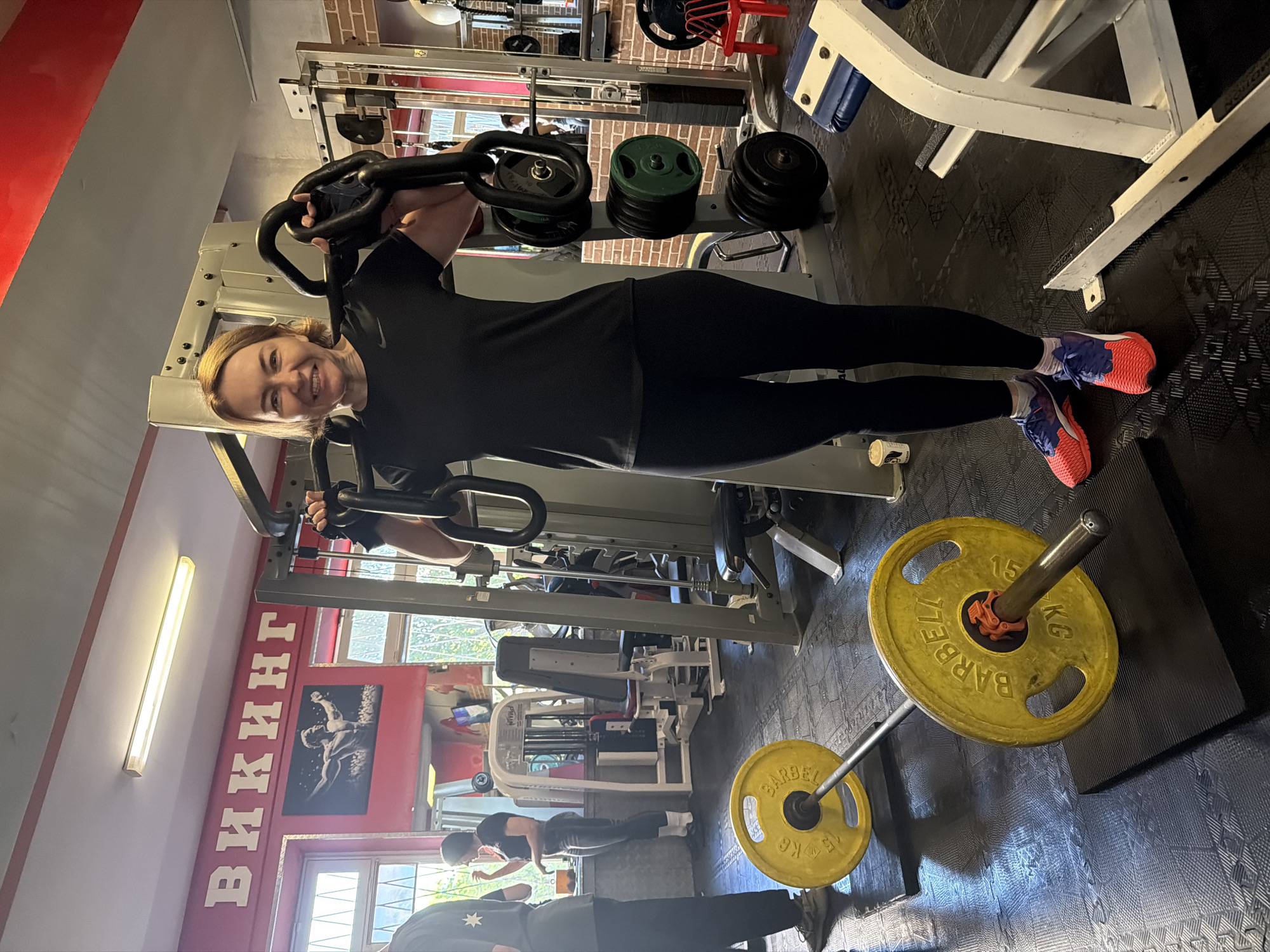 An image displays a woman smiling happily while exercising in a gym. She stands upright, facing forward, dressed in a black athletic top, black leggings, and distinctive coral and purple running shoes. Her light brown hair is pulled back, and she holds the handles of a standing gym machine, appearing to be in the middle of a workout, possibly an abdominal or leg raise exercise.
The gym environment features a combination of red-painted and exposed brick walls, with large windows allowing natural daylight to illuminate the space. The floor is covered with black, textured gym mats. Various pieces of fitness equipment are scattered around, including weight machines, weight stacks, and a white exercise bench. In the background, another person is partially visible, also engaged in exercise.
In the foreground, a barbell with two bright yellow 15 kg weight plates rests on the floor. The plates clearly display the text "BARBELL" and "15 KG". A prominent feature on the red wall is the word "ВИКИНГ" (Viking) written in large white letters, indicating the name of the gym. Given the location context, this gym is located in Saint Petersburg, Russia. The overall atmosphere appears active and positive, highlighted by the main subject's joyful expression.