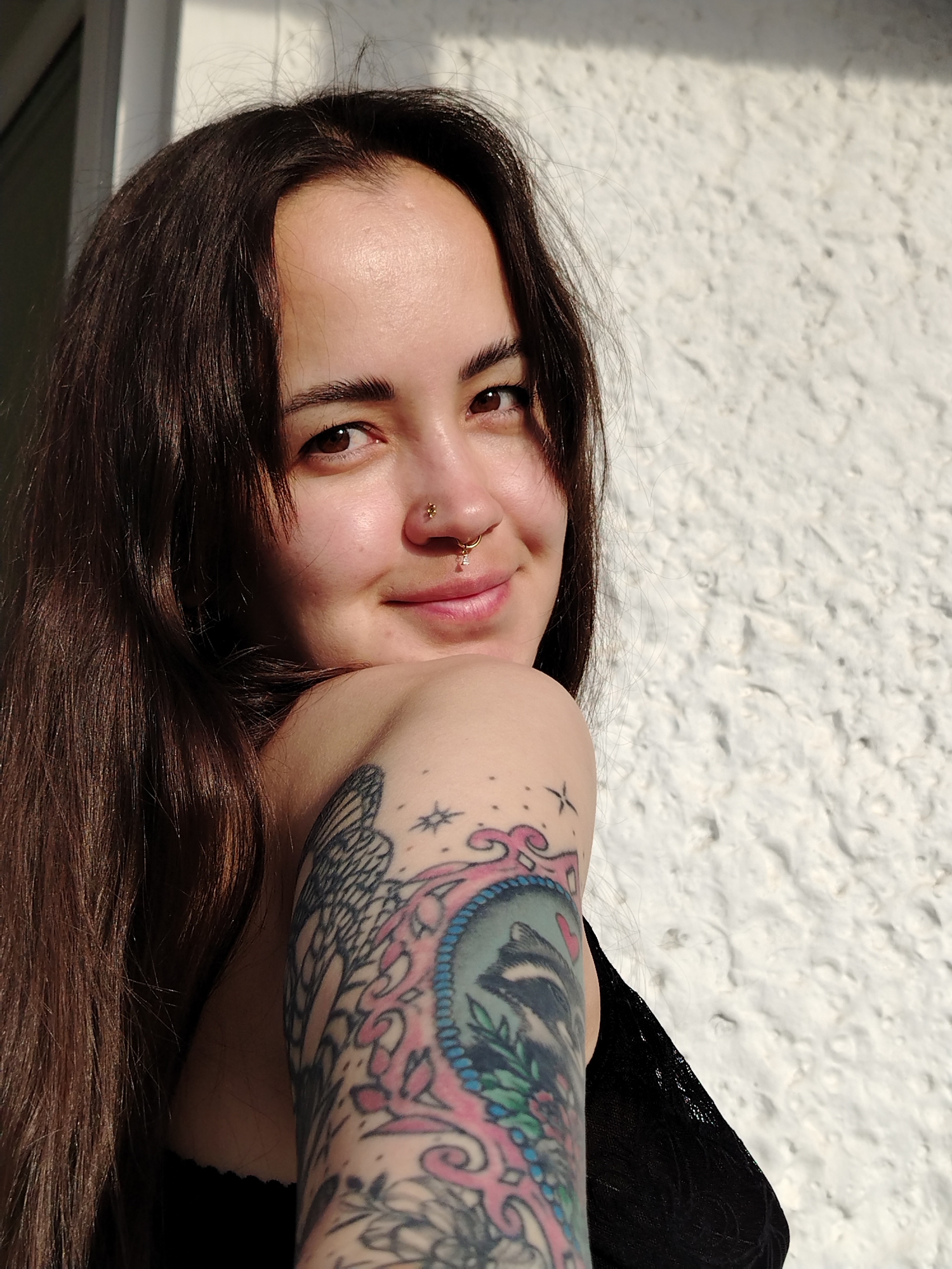 A close-up portrait captures a young woman with long, dark brown hair and brown eyes, looking over her left shoulder directly at the camera with a gentle, slight smile. She has a small gold stud piercing in her left nostril and a small hoop with a pink bead or charm adorning her philtrum. Her left upper arm is covered by a colorful, intricate tattoo featuring a raccoon's head enclosed in a decorative frame, surrounded by floral motifs and a large butterfly wing. She is wearing what appears to be a black top with some lace detailing visible on her shoulder. The background consists of a textured, off-white wall, brightly illuminated by direct sunlight from the right, creating distinct shadows and highlights. The strong lighting suggests it is a sunny day. There is no visible text in the image. The scene is set in Noviy, Russia.
