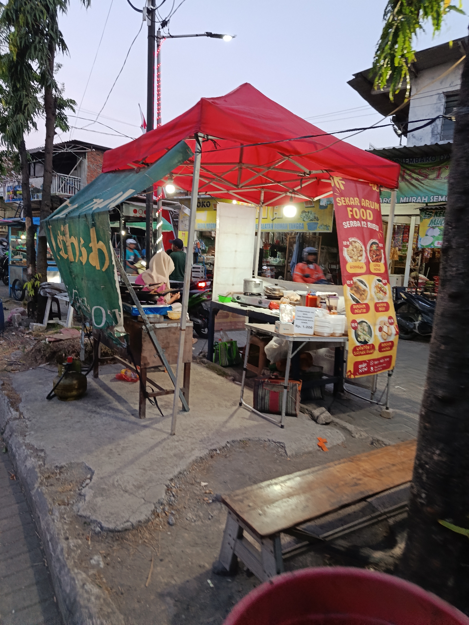 A street food stall with a red awning and a green tarp is set up on a concrete sidewalk in Keboansikep, Indonesia. A wooden table with stacked white containers and a red condiment bottle is positioned in front of the stall. Behind the table, a person wearing an orange jacket is visible, seemingly preparing food. A yellow banner with Indonesian text and images of food items is displayed vertically near the stall. A gas cylinder and various bags are placed on the ground. In the background, there are other stalls, buildings, trees, and a street with a parked motorcycle. The scene appears to be in the late afternoon or early evening, as indicated by the lighting conditions.