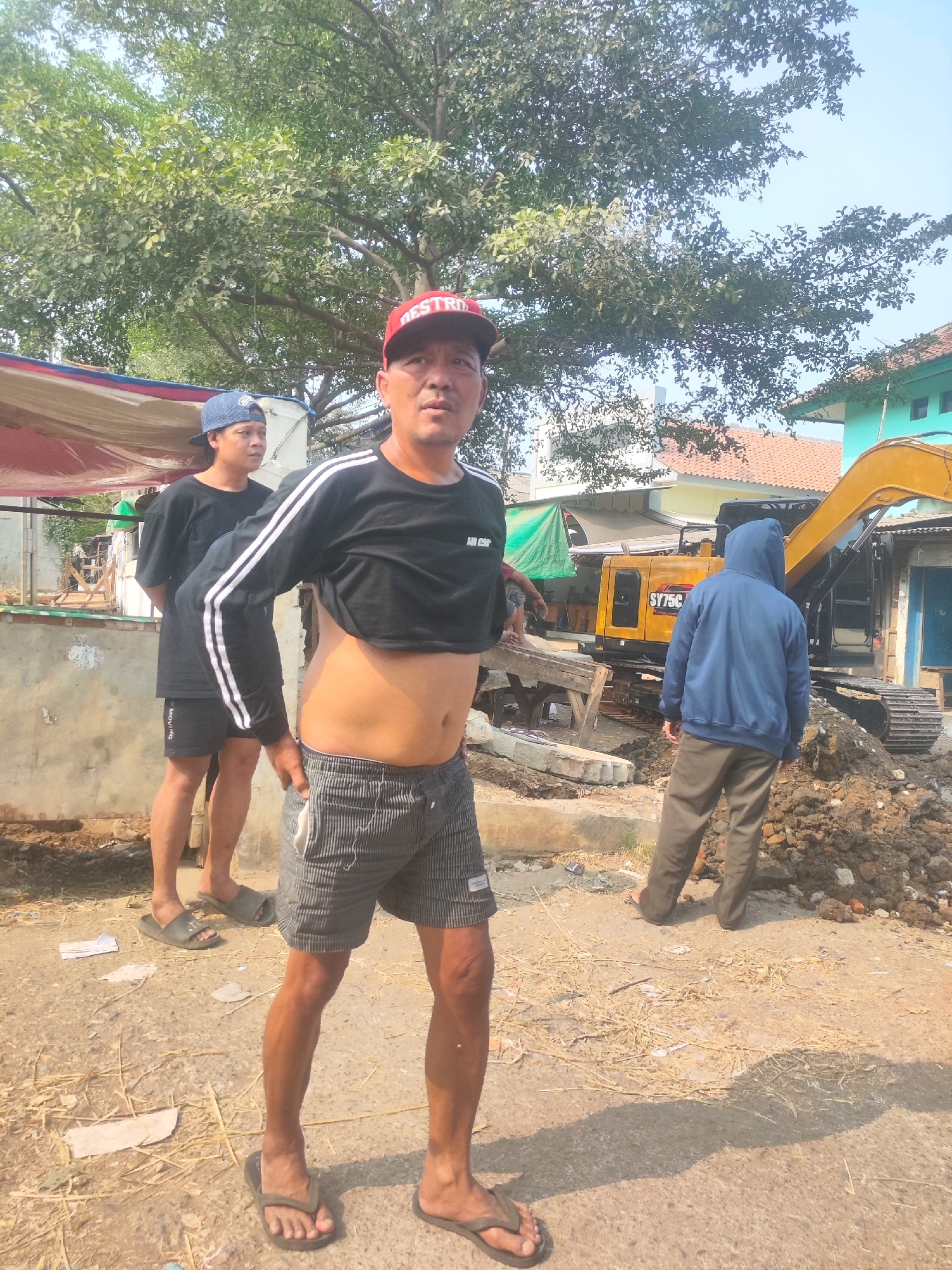 A low-angle medium shot captures three men standing in an outdoor area, likely a construction or repair site in Karawang, Indonesia. The central figure, a man wearing a red baseball cap and a cropped black long-sleeved shirt, faces forward with a neutral expression. His stomach is exposed, and he wears striped shorts and flip-flops. To his left, another man in a blue baseball cap and black t-shirt stands with his hands behind his back, looking to the left. To the far right, a third man in a blue hooded sweatshirt and brown pants is seen from behind, facing a yellow excavator. The background reveals a construction site with piles of dirt, wooden planks, and the excavator, suggesting ongoing work. The environment is characterized by dry ground, scattered debris, and overhead tree branches. The sky is a clear blue.