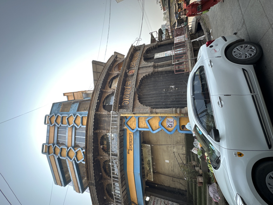 A building with ornate carvings and arched doorways stands adjacent to a street scene. The building's facade features a combination of blue and gold trim. A white car is parked in front, partly obscuring the view. The street appears busy, with pedestrians visible in the background. The image is taken from a slightly low angle, looking upwards towards the building.