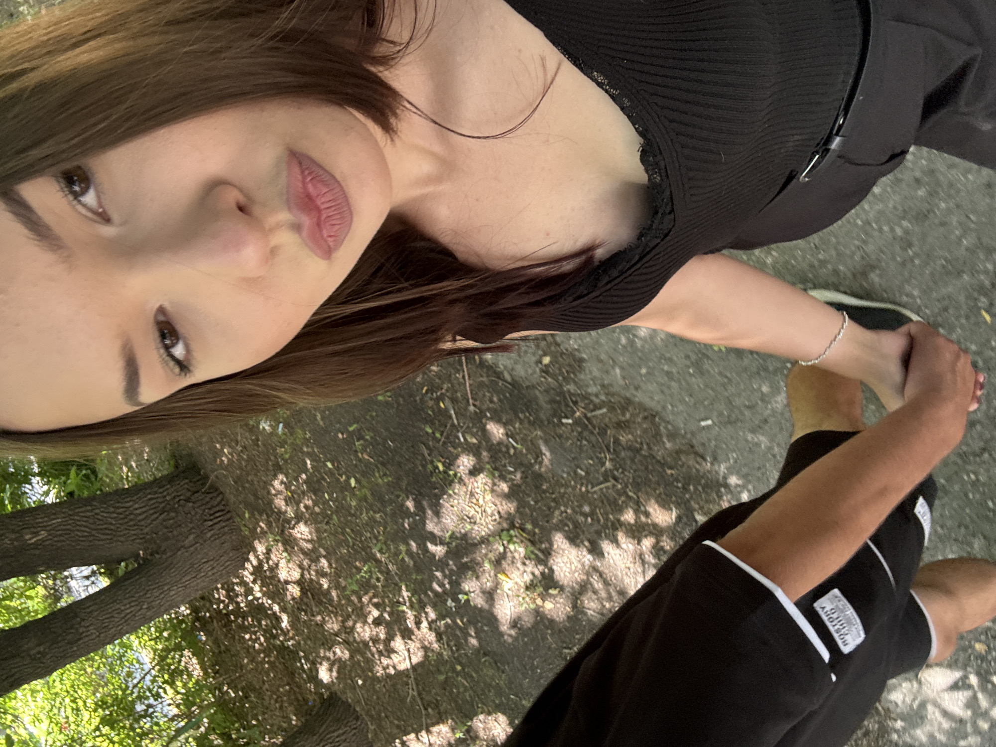 This is a selfie taken outdoors, likely in a park or wooded area. In the foreground, a woman is holding hands with someone whose arm and legs are visible on the left side of the frame. The woman is wearing a black sleeveless top and appears to be looking at the camera, with her lips slightly parted. The person on the left is wearing black shorts with white trim and a white tag that reads "ROSTORY CHILD". The ground is a mix of dirt and pavement, with fallen leaves and dappled sunlight creating shadows. In the background, trees with rough bark are visible, suggesting a natural, outdoor setting. The presence of trees, the lighting, and the overall atmosphere are consistent with a park or a natural area within a city, such as in Belovo, Russia.