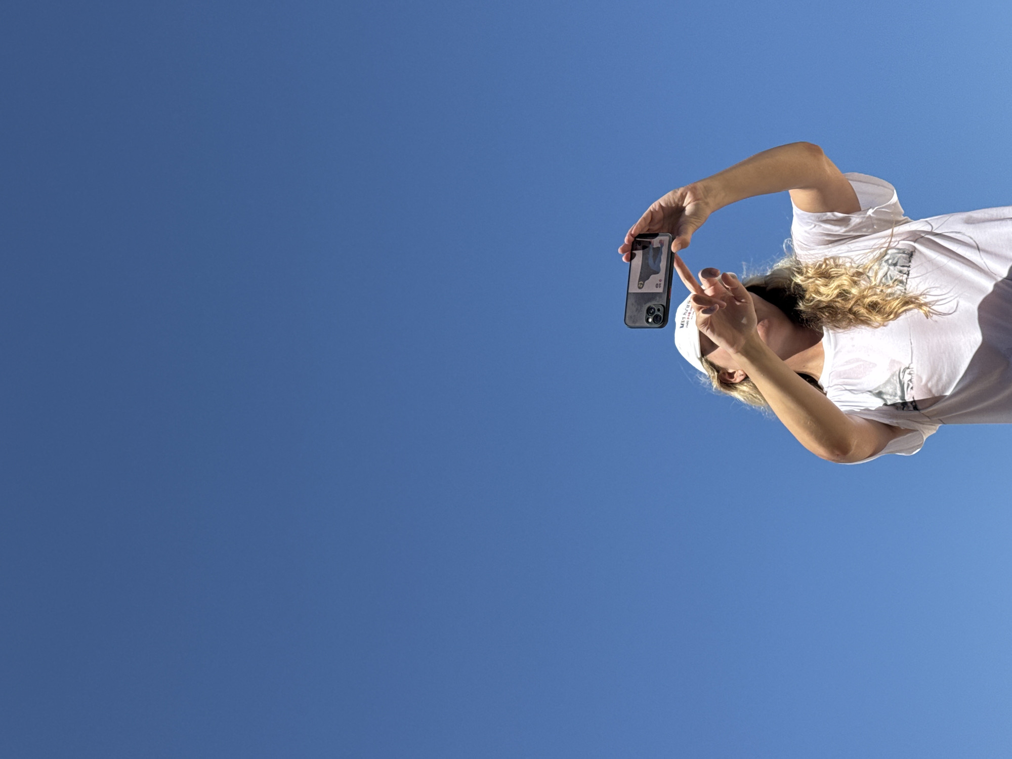 A woman wearing a white t-shirt and white cap, with blonde curly hair, is holding a smartphone horizontally to take a picture of herself against a clear blue sky. Her left hand is visible, holding the phone, while her right hand is partially covering her face, with her fingers extended. The phone screen displays an image. The background is an expanse of clear blue sky. The scene is not specifically identifiable as being in Kuta Selatan, Indonesia, but the bright, sunny sky and beach-appropriate attire could be consistent with such a location.