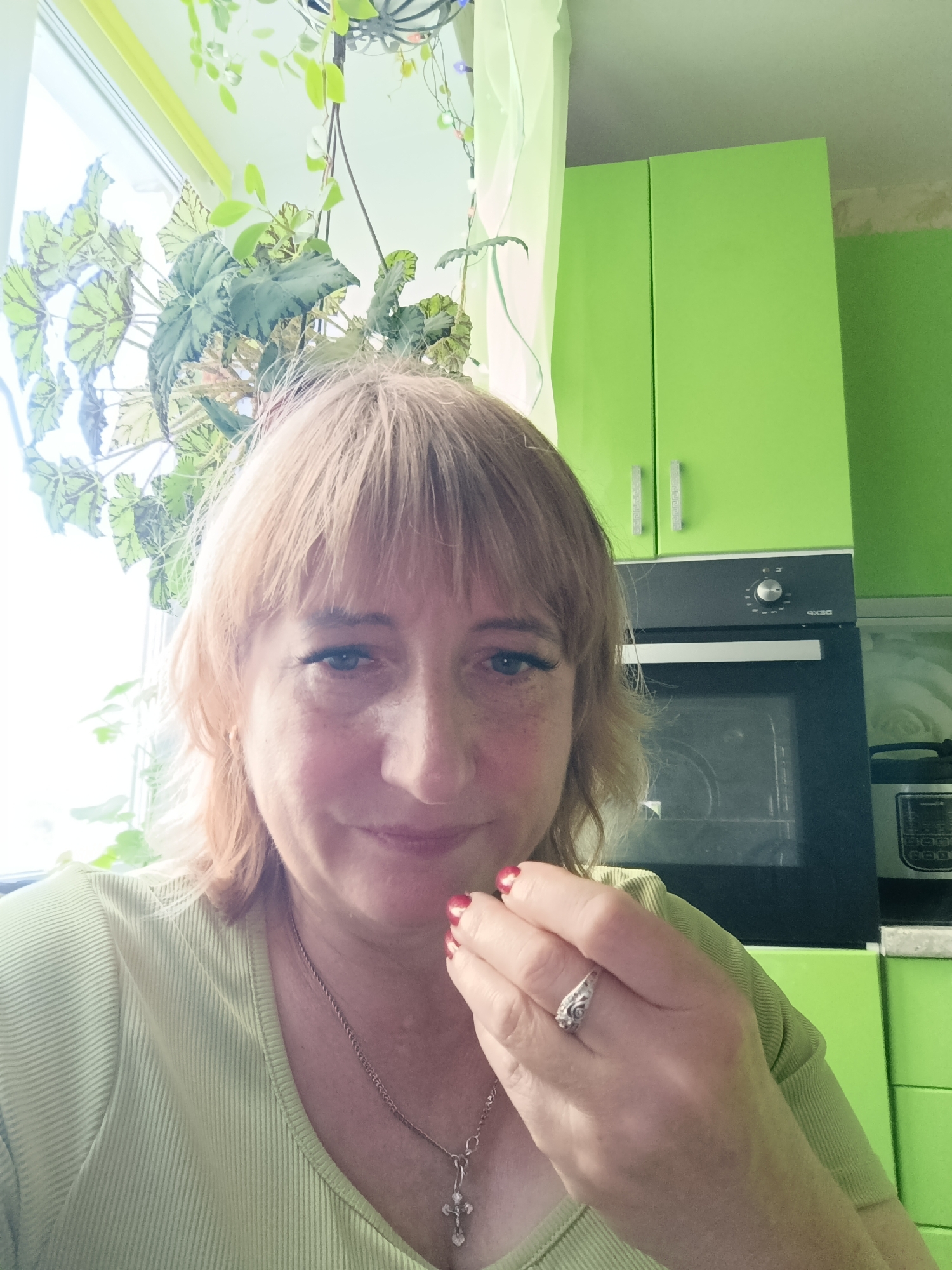 A woman with shoulder-length blonde hair and bangs, wearing a light green ribbed top and a silver necklace with a cross pendant, is captured in a close-up, selfie-style portrait. She has blue eyes, light skin with freckles, and is wearing makeup, including mascara and possibly eyeliner, accentuating her eyes. Her right hand, with red-painted nails and a diamond ring on her left ring finger, is raised to her face, near her mouth. She has a slight smile, conveying a sense of warmth or slight amusement.
The background suggests an indoor setting, likely a kitchen, with bright lime green cabinets visible. An oven is also present, integrated into the cabinetry. To the left of the woman, a window is partially visible, allowing natural light to enter and illuminating a cluster of lush green plants hanging from above. The plants appear to be trailing or vine-like with broad, patterned leaves. The lighting suggests it might be daytime, possibly morning or afternoon, given the natural light from the window. There is no visible text in the image. The overall impression is a casual, everyday moment captured at home.
