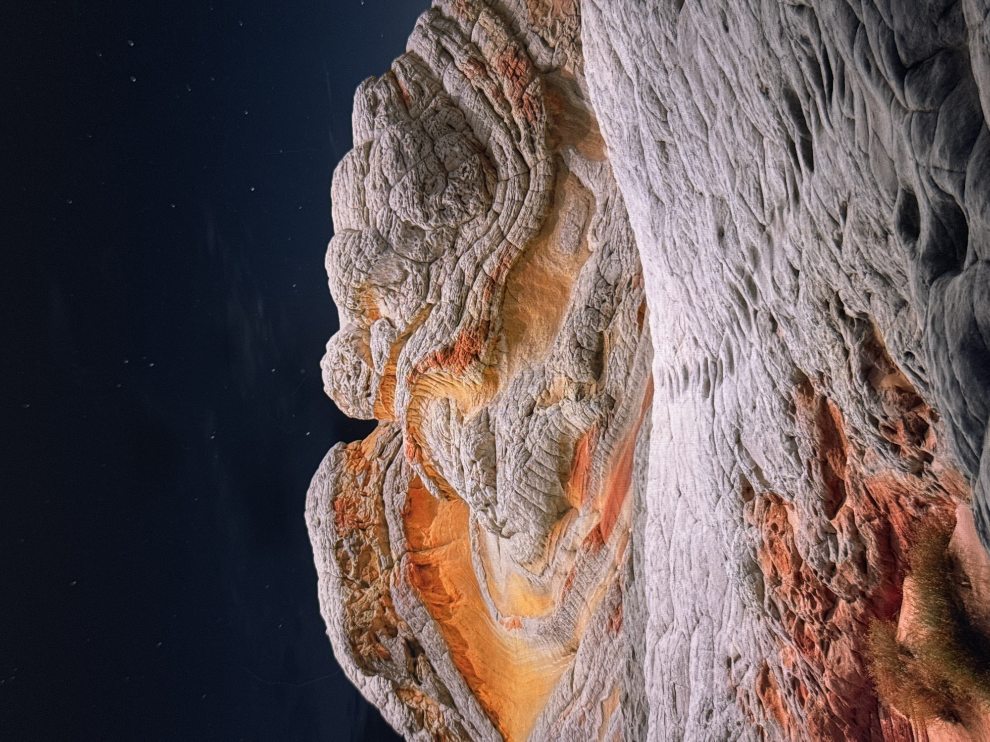 The image shows a close-up of layered rock formations. The rock exhibits a range of colors, from light gray to orange and reddish-brown. The formations appear to be eroded, with distinct patterns and textures. The background is dark, resembling a night sky with small points of light that could be stars or dust. There is no evidence to suggest the image is located in Riga, Latvia, or any other urban location. There are no people, subjects, objects, actions, or interactions visible in the environment.