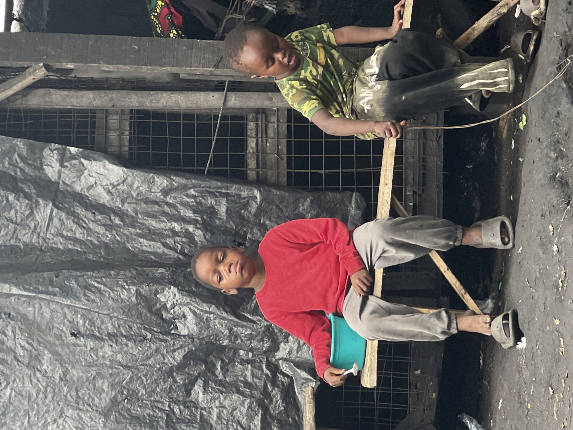 Two young boys are pictured outdoors in what appears to be an informal settlement in Nkpor, Nigeria. The scene is somewhat cluttered and appears to be in an area with rough ground and rudimentary structures.
The boy in the foreground is wearing a red long-sleeved shirt and grey sweatpants, sitting on a wooden plank. He is holding a teal-colored bucket with his left hand and a small white object, possibly a spoon, in his right hand. He is looking towards the camera with a neutral expression. His feet are bare except for grey flip-flops.
The second boy, positioned higher and further back, is wearing a camouflage t-shirt and dark pants with white stripe accents. He is also sitting on wooden planks. His eyes are closed, and he appears to be resting or sleeping. He is leaning against a wooden structure.
The background consists of a dark, rough wall or corrugated metal sheeting, and a wire mesh fence. A large piece of black plastic sheeting is visible in the foreground and middle ground, creating a backdrop for the boy in red. A piece of patterned fabric with red, yellow, and black colors is visible behind the boy in the camouflage shirt, possibly part of clothing or a blanket.
The lighting suggests it is daytime, but the overall environment appears somewhat dim. The ground is dirty and dusty. There is no clear indication of specific weather conditions. The overall impression is one of a humble and possibly challenging living environment. There is no visible text in the image.