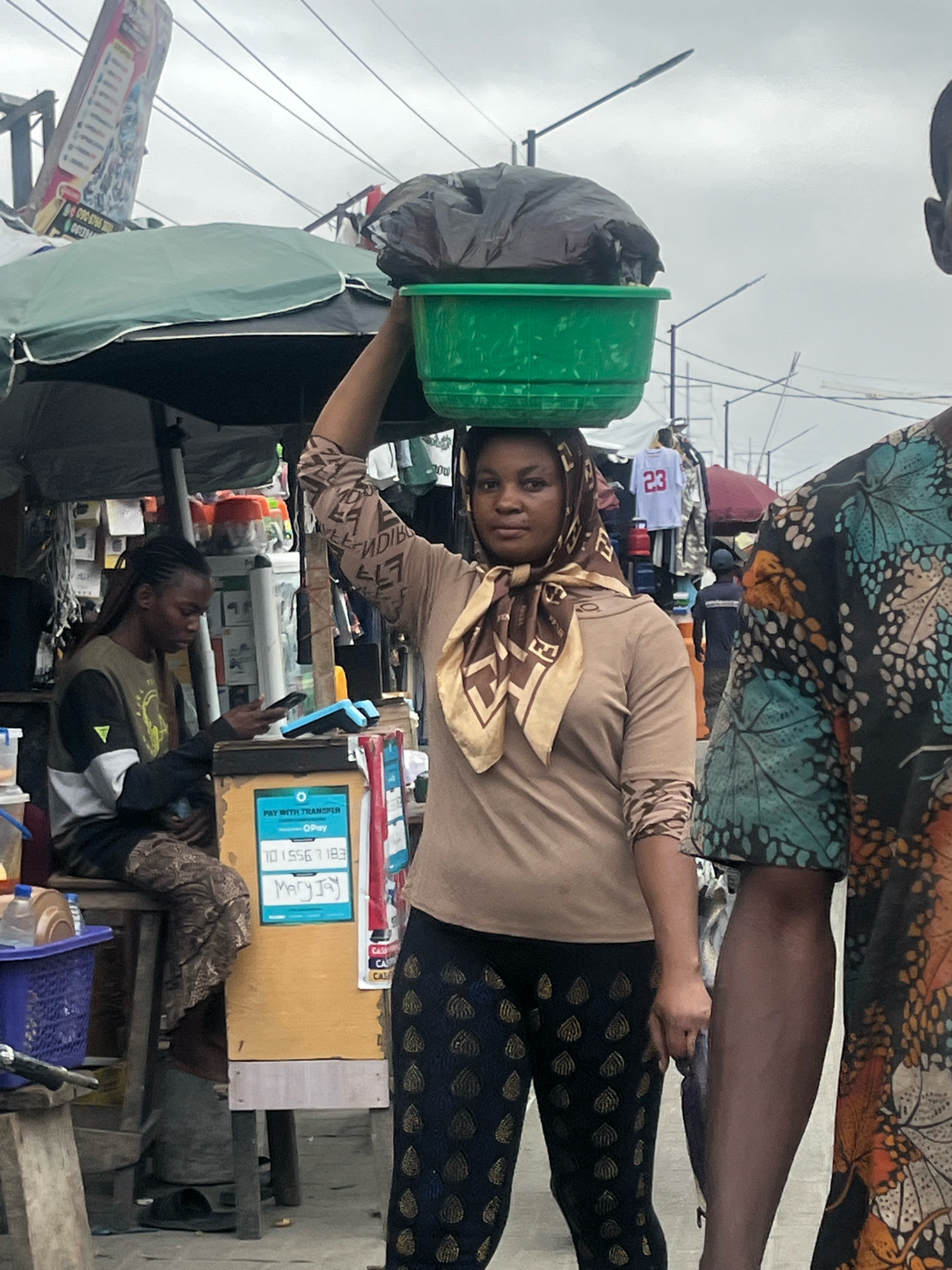 A street scene in Lekki Phase I, Nigeria, features a woman balancing a green plastic basin filled with a black plastic bag on her head. She is wearing a tan long-sleeved shirt with a patterned scarf around her neck and dark trousers with a repeating gold leaf pattern. To her left, a person is seated on a stool, looking at a mobile phone and interacting with a point-of-sale device. This individual wears a long-sleeved shirt with contrasting black, white, and olive green sections. In the foreground on the right, a person's arm and torso are visible, clad in a black shirt with a vibrant, multi-colored butterfly or leaf pattern. The background shows market stalls with various items for sale, including clothing and electronics, under a cloudy sky. Overhead, power lines and streetlights are present.