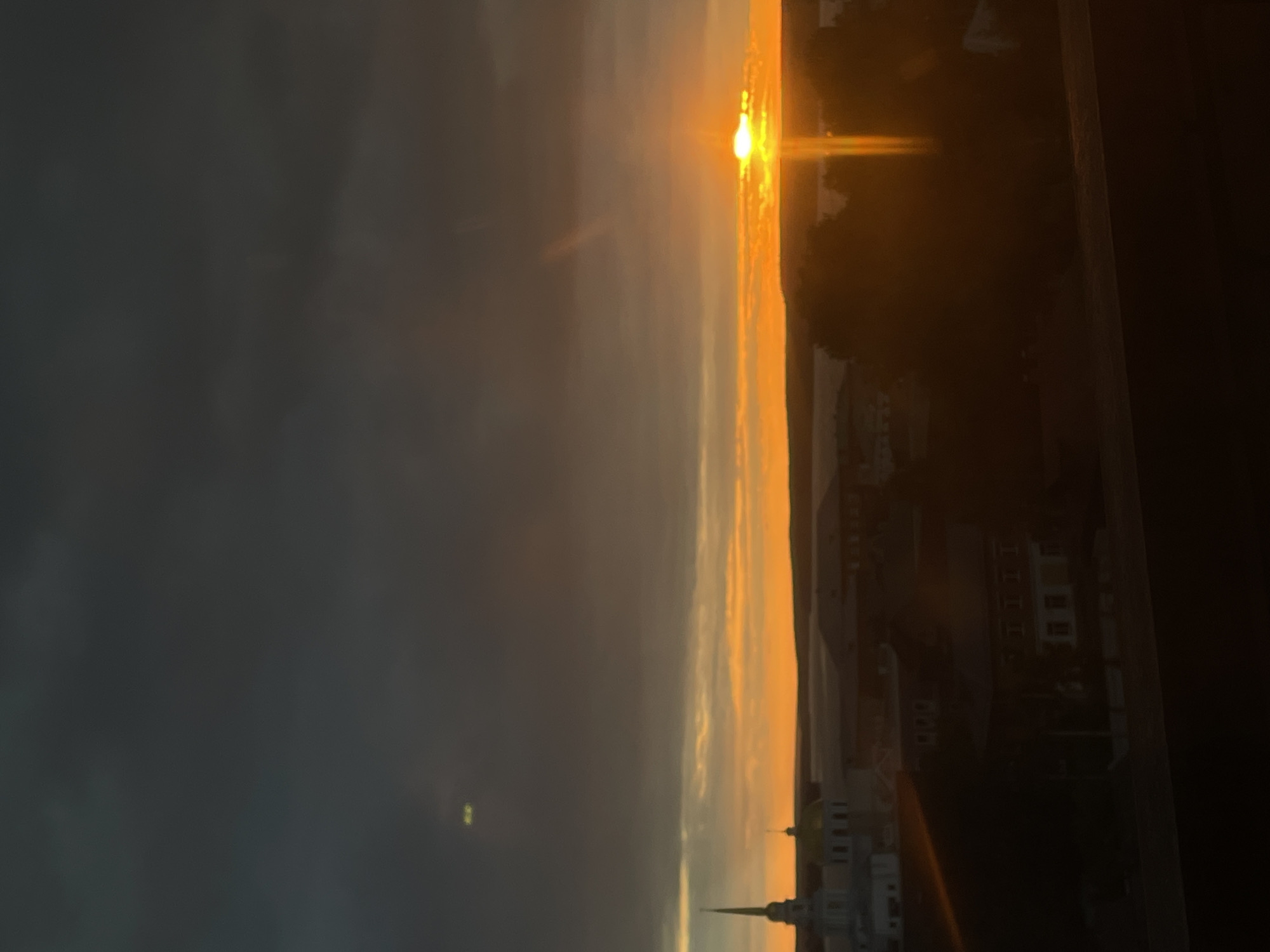 This vertical, full-frame, eye-level shot captures a sunset over the city of Votkinsk, Russia. The sky is dominated by dark, ominous clouds, with a sliver of bright orange light breaking through on the horizon where the sun is setting. The sun itself is a small, intense point of light, creating a strong lens flare that extends upwards.
In the middle ground, the silhouette of Votkinsk is visible. A prominent church with a tall, slender spire and a golden dome stands out on the left. To the right of the church, there are other buildings and trees, all silhouetted against the vibrant sunset. The foreground is a dark, blurred area, possibly a railing or barrier, adding to the sense of depth. The overall mood is dramatic and somewhat melancholic due to the dark clouds contrasting with the brilliant sunset.