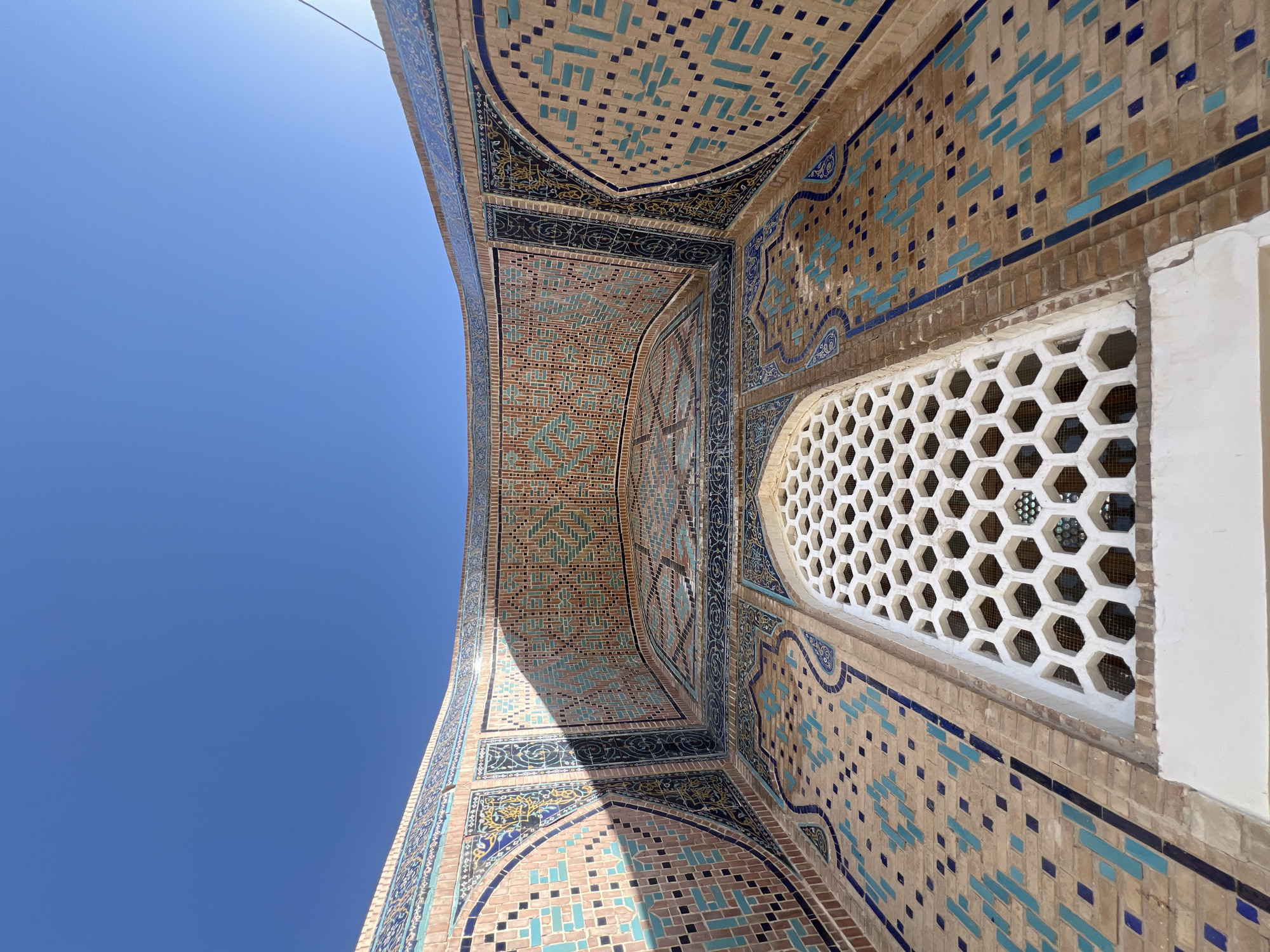 This image shows a low-angle shot of a decorative facade on a building, likely a religious or historical site, under a clear blue sky. The facade features intricate tilework in shades of blue, turquoise, and terracotta, forming geometric patterns. A large, hexagonal lattice window is prominently visible in the lower center of the image. The building's architecture suggests it is located in Central Asia, possibly in Samarkand, Uzbekistan, known for its rich Islamic architecture. The lighting indicates a bright, sunny day, casting shadows on the tiled surfaces. There are no people or animals visible in the frame. The overall impression is one of architectural beauty and cultural heritage.