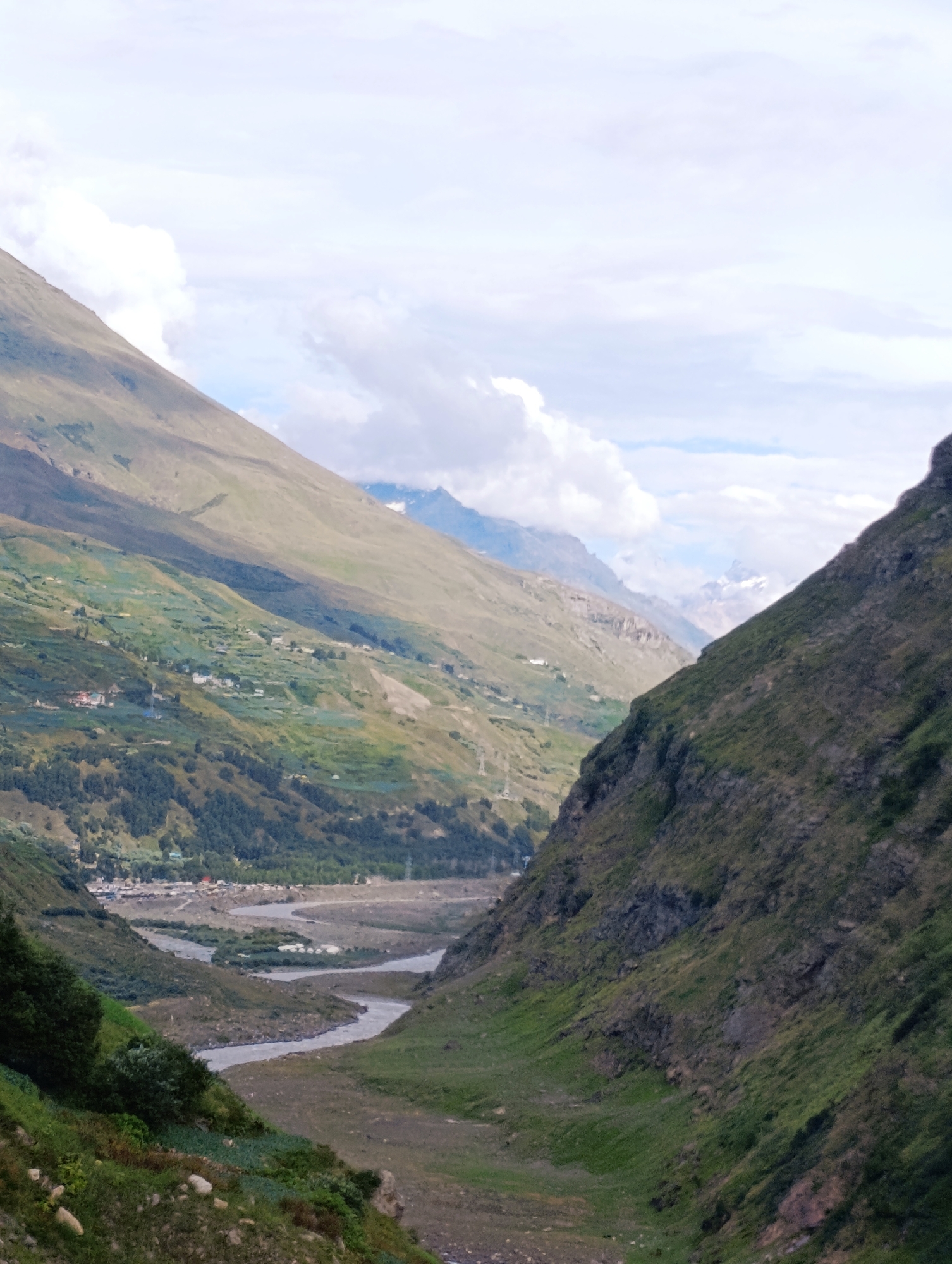 An aerial, vertical shot captures a wide valley in Keylong, India, during the daytime. Steep, verdant mountains flank the valley on both sides, with a winding, grey river flowing through its center. The slopes are partially cultivated, showing patches of green crops and some scattered white buildings that form a small village. In the distance, further mountains are visible, some with snow-capped peaks and a clear blue sky with scattered clouds above. The overall scene is one of natural beauty and rural tranquility.