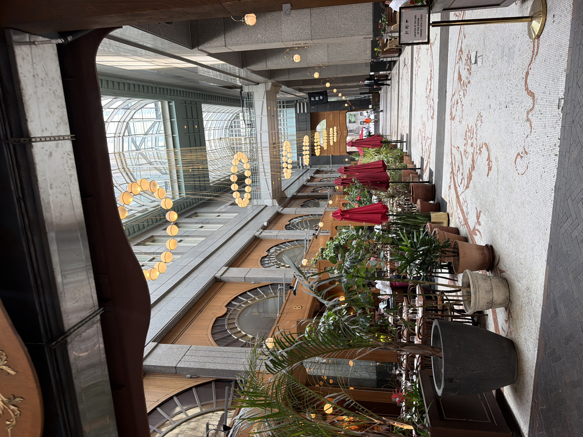 The image depicts an interior view of a grand atrium or shopping arcade, likely in New York City, given the architectural style and commonality of such spaces in the city. The perspective is looking down the length of the atrium.
On the left side of the foreground, a section of an outdoor seating area for a restaurant is visible. This includes several large potted plants, notably a tall palm tree in a dark gray planter, and various smaller plants. Red umbrellas are set up over tables and chairs, some of which are set with glassware. A person, presumably a server or staff member, is visible behind the tables.
The atrium itself is characterized by a mosaic tiled floor with intricate reddish-brown patterns. The walls of the atrium are constructed of granite and wood paneling, with large arched windows featuring decorative glasswork.
Above, the atrium ceiling is a glass structure, allowing natural light to filter in. Suspended from the ceiling are multiple tiers of decorative light fixtures, resembling circular chandeliers composed of many small, round bulbs. A walkway or bridge spans the atrium at a higher level, connecting the buildings on either side.
Down the length of the atrium, several individuals are walking, including a man in a dark suit and tie on the right side. On the right wall, a sign is mounted on a brass stand. The sign has Japanese characters at the top and English text below, stating "LOCATED AT THE END OF THE ATRIUM TOWARD 54TH STREET". This text suggests the location is within a building with access to 54th Street.
The overall impression is one of a sophisticated and well-maintained urban commercial space, likely a high-end shopping center or a building with multiple businesses and restaurants.