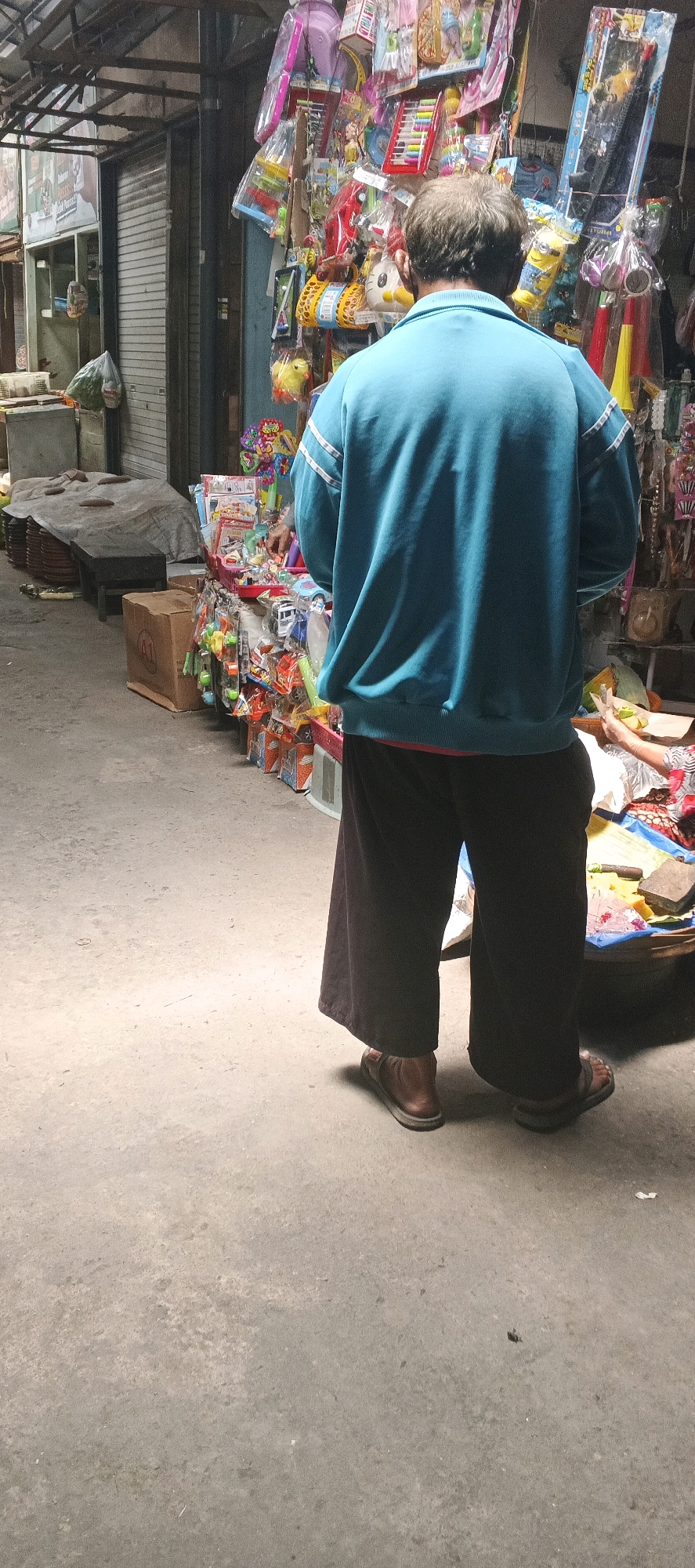 A person stands with their back to the camera in an outdoor marketplace. The individual is wearing a turquoise sweatshirt with white stripes on the sleeves and dark, wide-legged pants. They are also wearing brown sandals. The person is facing towards a stall filled with various toys and novelty items, some of which are still in their packaging. To the left of the frame, there are closed shopfronts with metal shutters and more stalls displaying goods. The ground is a concrete surface. The overall scene depicts a busy marketplace in Maduran, Indonesia.