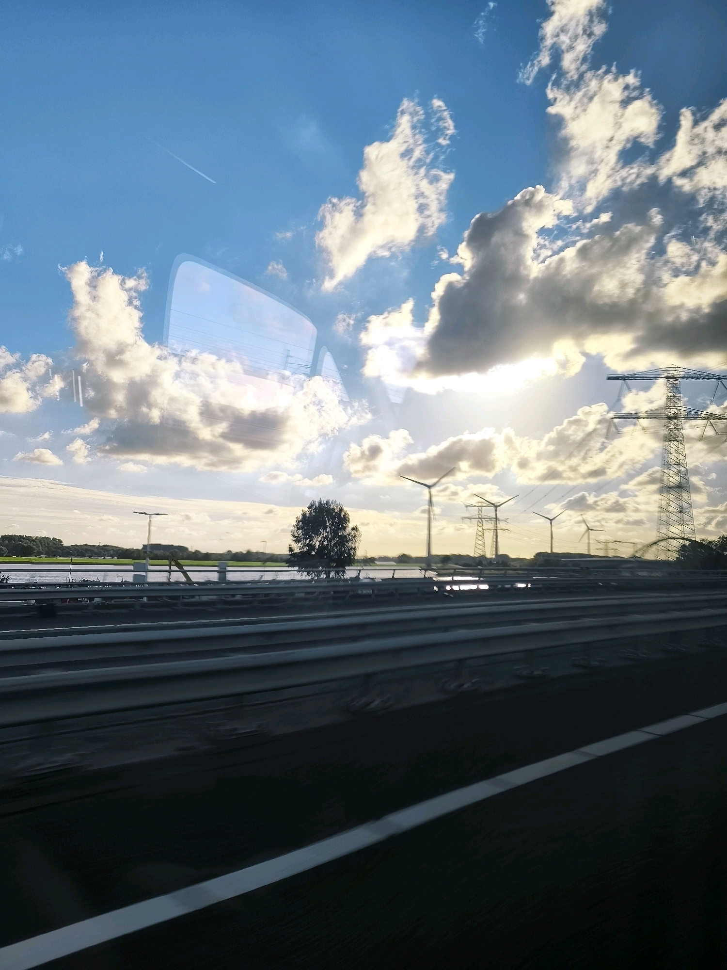 The image depicts a view from a vehicle traveling on a highway in Europoort Rotterdam, Netherlands. The scene is dominated by a bright, cloudy sky with the sun partially obscured by clouds, casting a warm glow. In the distance, there are several wind turbines and large electricity pylons, indicative of the area's industrial and energy production landscape. A body of water, likely a canal or river, runs parallel to the highway. A single tree stands on the bank of the water. The highway itself is visible in the foreground with distinct lane markings. There are reflections on the vehicle's window, suggesting the photograph was taken from inside a moving vehicle. No people are visible in the frame. The overall atmosphere is one of a bright, possibly late afternoon or early morning, sky over a functional, industrial environment.