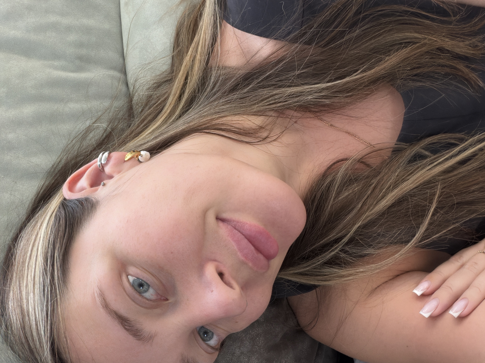 A fair-skinned woman with light blue eyes and long, highlighted brown hair is reclining on a gray upholstered surface. She has multiple piercings in her left ear, including a double helix piercing with silver rings and a stud earring. She is wearing a black top and a thin gold chain necklace. Her nails are manicured with a French tip style. The background is out of focus, suggesting an indoor environment. The location is not discernible from the image.