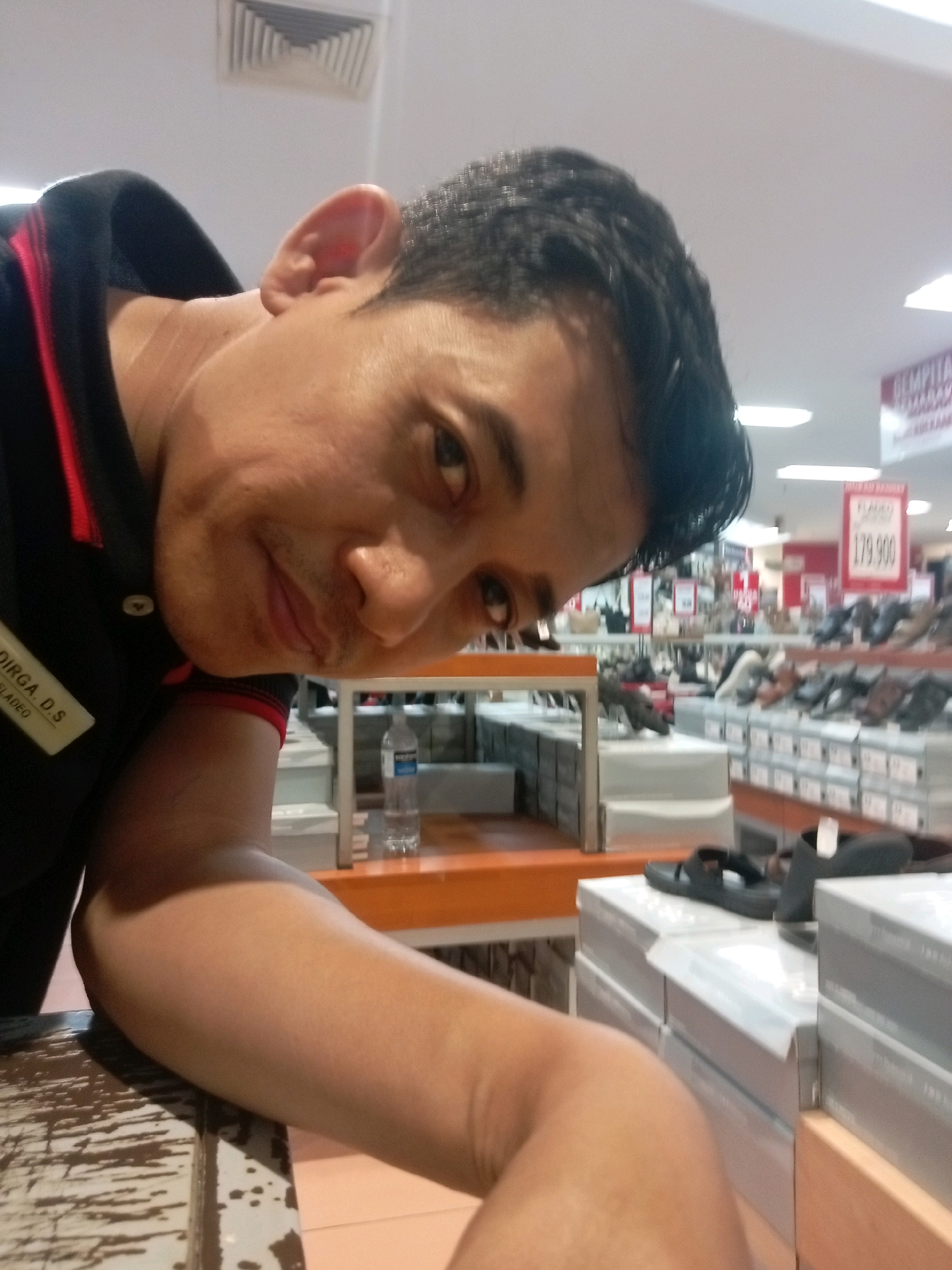 A man is taking a selfie in what appears to be a shoe store. He is wearing a black shirt with red trim. The background shows shelves filled with shoe boxes and a sign with a price of 179,900. The lighting suggests an indoor commercial setting. The man is looking directly at the camera, and his right arm is resting on a table or counter.
