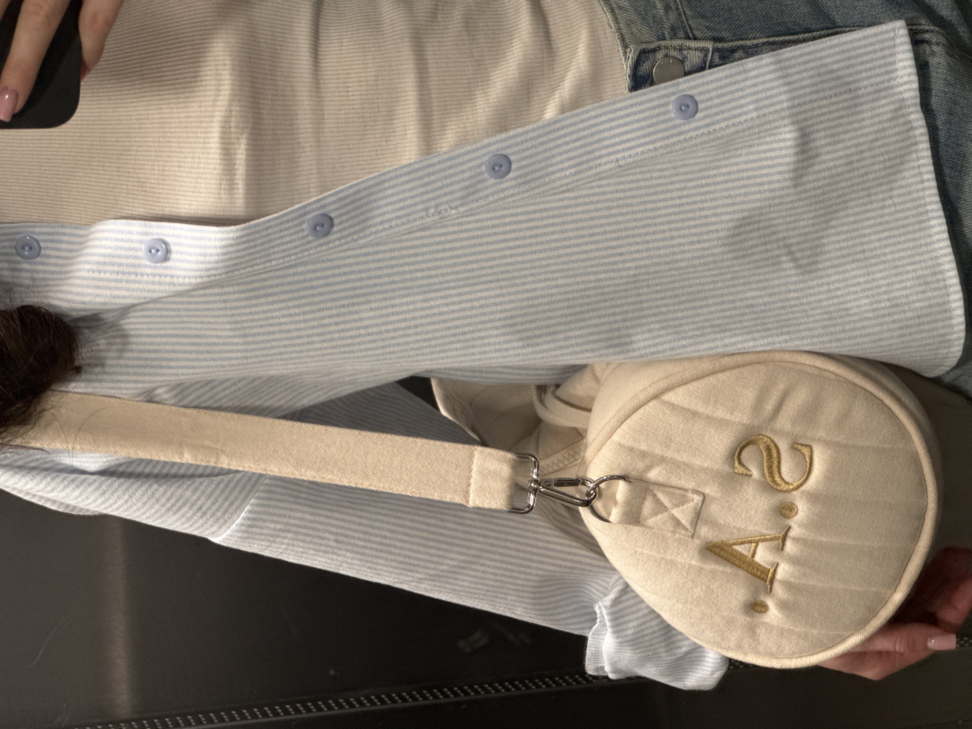 The image shows a person wearing a light blue and white striped button-down shirt. The shirt appears to be part of an outfit that includes a cream-colored tank top and a denim jacket. A small, cylindrical cream-colored bag with gold embroidered letters "2.A." is positioned on the person's shoulder, held by a cream-colored strap with a metal clasp. The person is holding a phone, and their manicured fingers are visible. The background is dark and unclear. The location is not identifiable as Unknown, Unknown based on the image.
