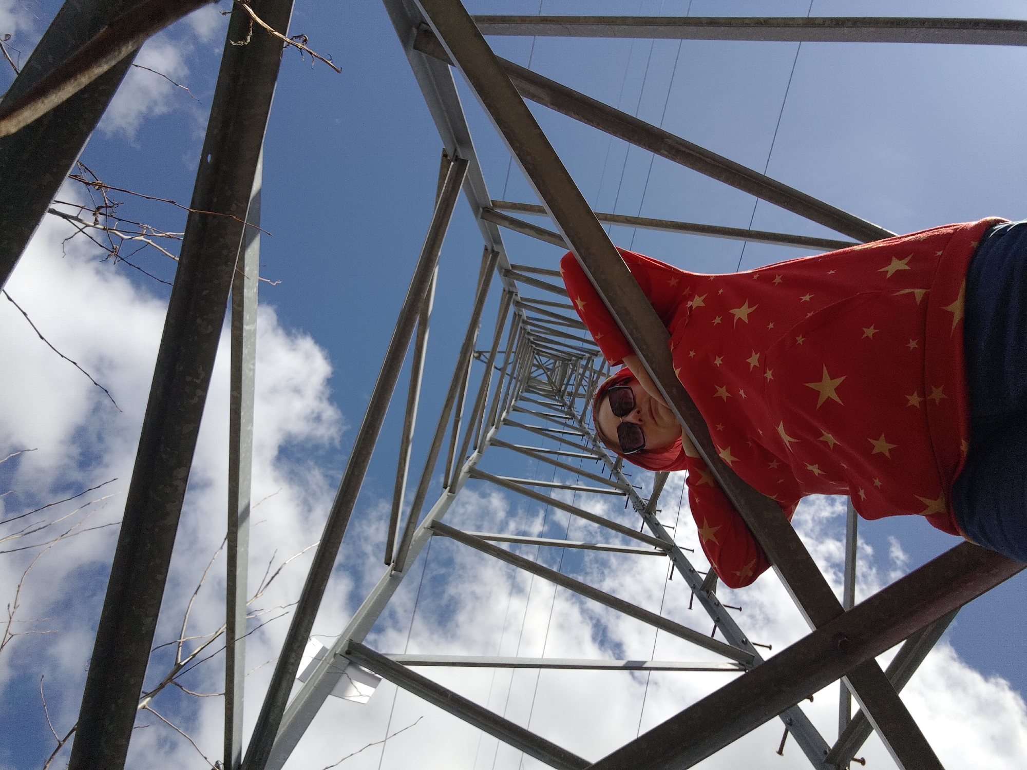 A low-angle shot depicts a person with sunglasses wearing a red star-patterned hoodie, seemingly positioned within or alongside a metal power-line tower. The structure is composed of crisscrossing steel beams, angled sharply upwards. Behind the tower, a blue sky with scattered white clouds is visible. In the lower left corner, thin bare branches extend into the frame. The specific location in Chaikovsky, Russia cannot be determined from the image alone.