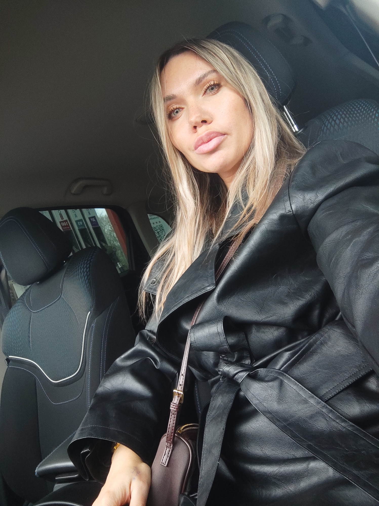 A fair-skinned woman with blonde hair sits in the driver's seat of a car. She wears a black leather jacket tied at the waist, and a brown purse is visible. Through the car window, one can see gas pumps in the city of Votkinsk, Russia.