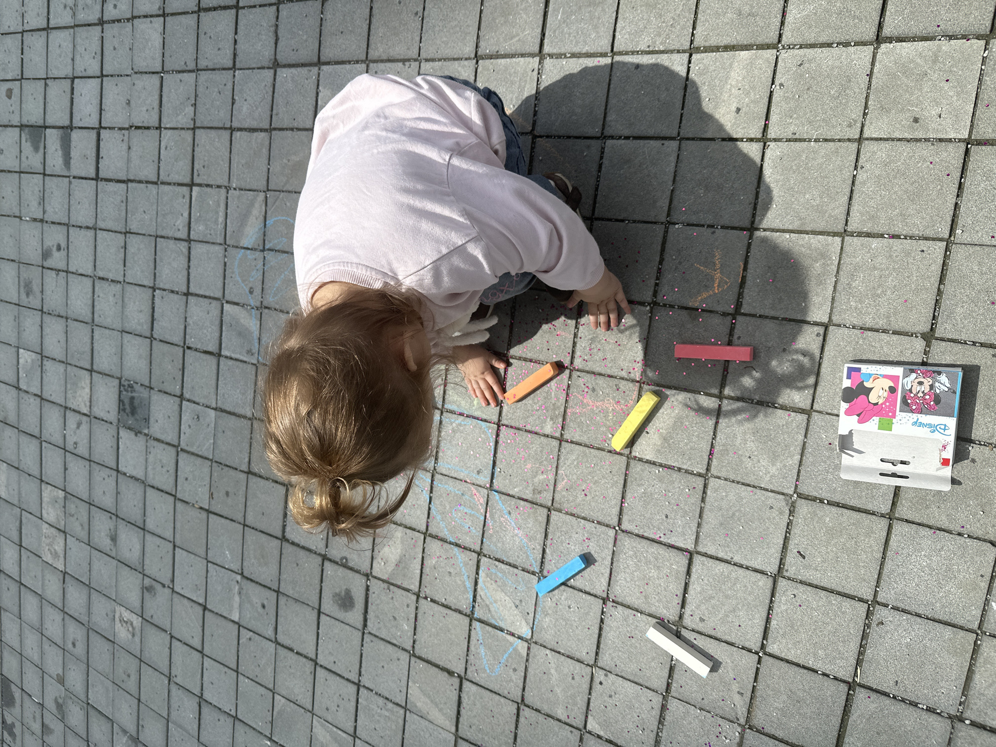 An overhead outdoor shot captures a young child, characterized by light brown hair tied in a top bun, wearing a light pink long-sleeved sweatshirt, blue jeans, and dark shoes, kneeling on a grey tiled pavement. The child's hands are positioned on the ground amidst scattered art supplies. Five pieces of chalk are visible: blue, orange, yellow, white, and red. A partially opened "Disney Minnie Mouse" branded chalk package rests in the bottom left. Numerous small, pink, heart-shaped glitter pieces are liberally scattered across the tiles, particularly concentrated around the child and the chalk. Faint chalk drawings are present on the pavement, including light blue outlines resembling an abstract shape or animal on the left, and a small orange mark near the red chalk. The ground consists of uniformly sized grey square concrete tiles arranged in a grid pattern. Strong sunlight casts distinct shadows of the child and chalk onto the pavement, indicating a bright daytime outdoor setting. No specific identifying markers for the city of Yekaterinburg, Russia, are discernible within the image.