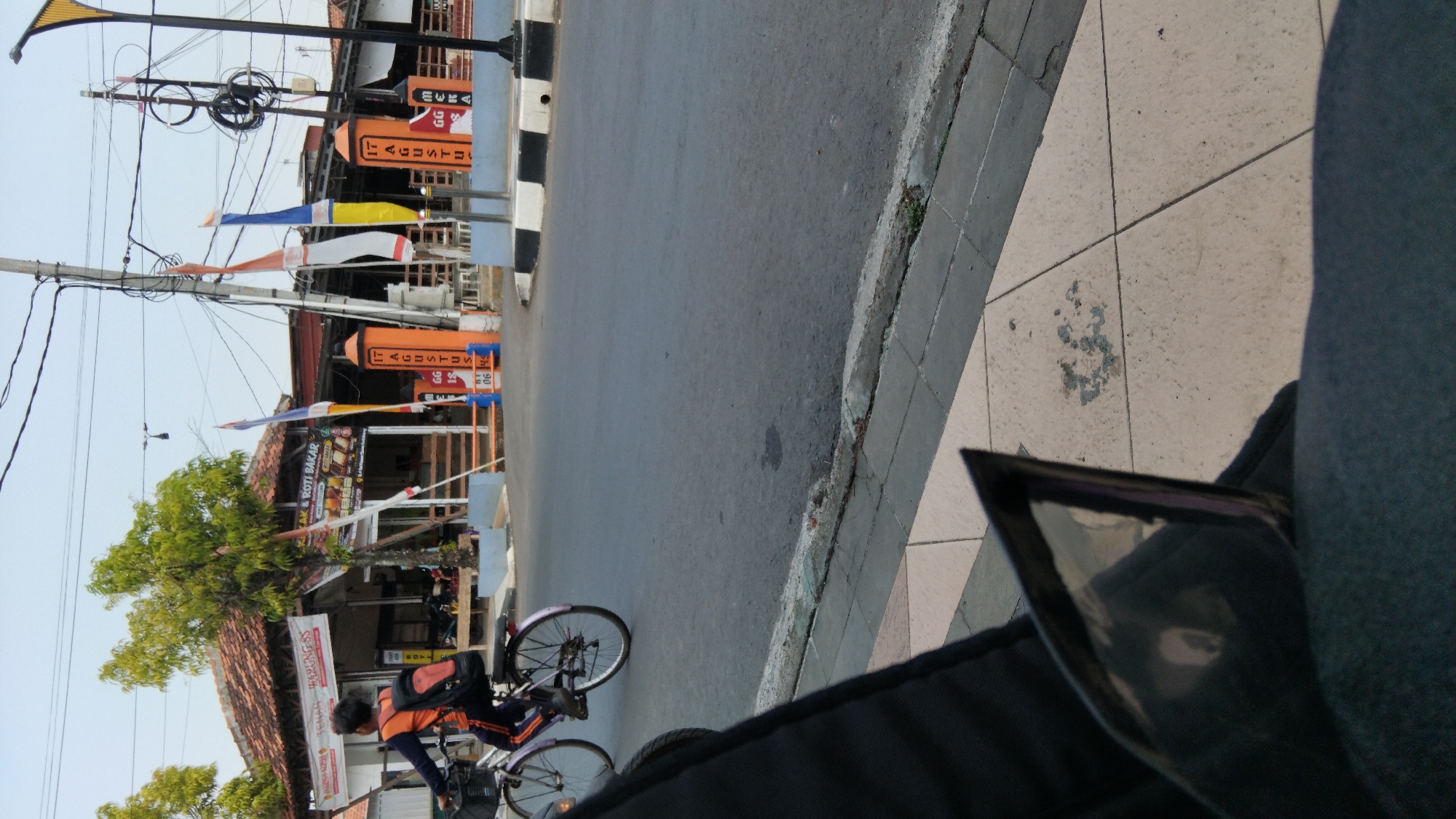In Batang, Indonesia, a person rides a bicycle on a street in front of a building with a red-tiled roof. The building appears to be a food establishment, with signs reading "MAKAN & ROTI BAKAR" (Eat & Grilled Bread) and other Indonesian text. Flags of red and white, along with yellow and blue flags, are displayed in front of the building. Traffic poles with electrical wires are visible overhead. The street is paved, with a sidewalk and a curb on the side. The foreground shows a partially visible dark object, possibly a piece of furniture or fabric, with a reflection on its surface.