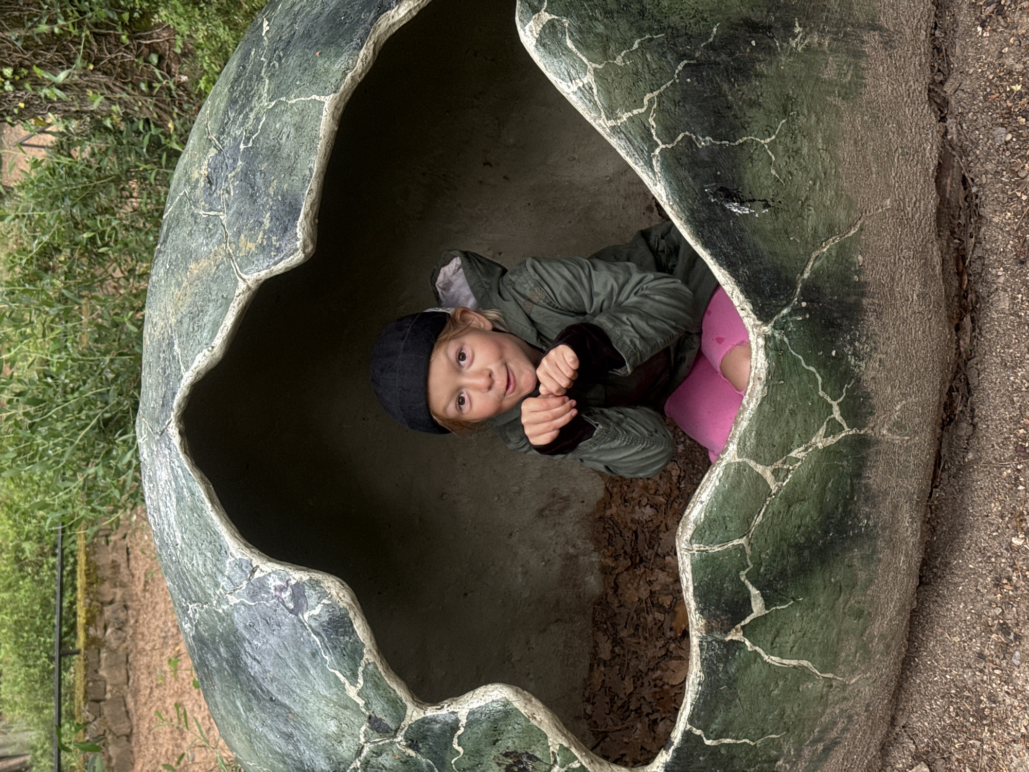 A child is inside a large, cracked egg-shaped structure. The child is wearing a black cap, a green jacket with brown sleeves, and pink leggings. They are looking up and making a gesture with their hands. The inside of the egg appears to be concrete or a similar material, with some leaves and debris on the bottom. The exterior of the egg is a greenish-gray color with white cracks running across the surface. The egg is situated outdoors, as evidenced by the gravelly ground to the right and vegetation visible in the background on the left. The location and city are unknown.