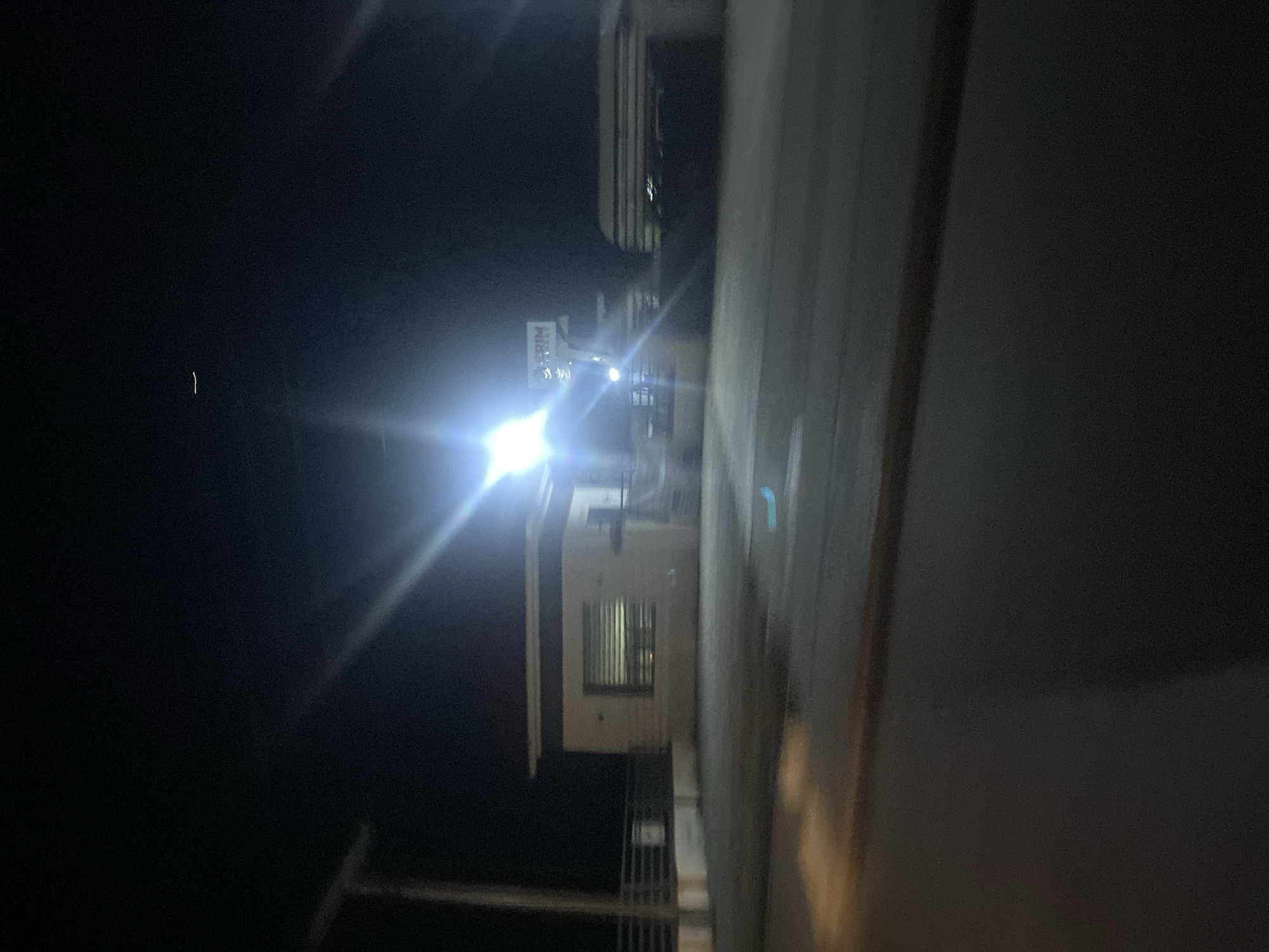 The image shows a security checkpoint at night in Jalingo, Nigeria. A small, white booth with a window is visible, and there appears to be a sign above it with the letters "TRIM". A bright light shines from the left, illuminating the scene and creating lens flare. In the background, there are some vehicles and structures. The ground is paved.