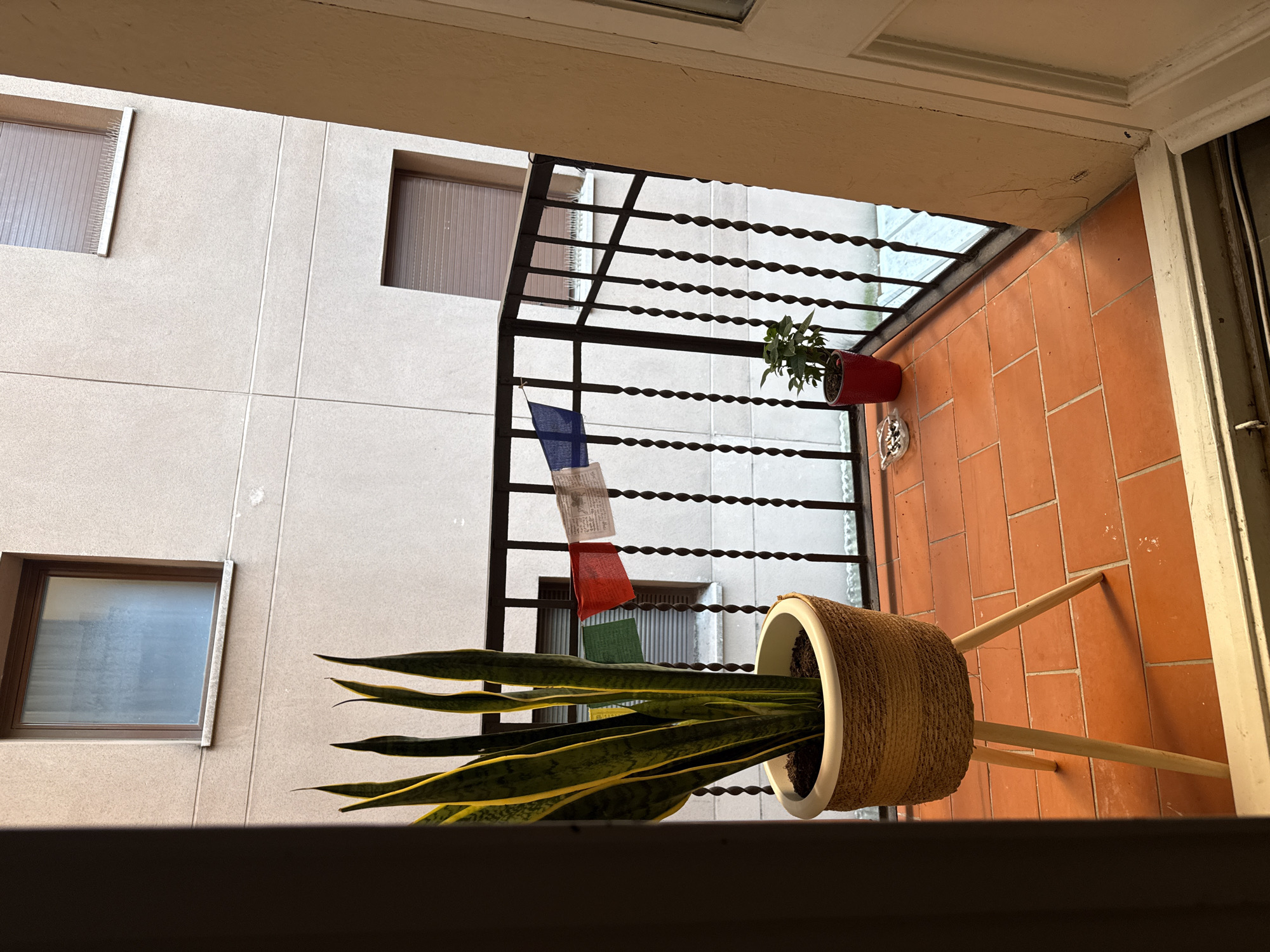 The image captures a balcony in a residential building in Barcelona, Spain. The balcony floor is tiled with orange terracotta. A black metal railing runs along the edge of the balcony, adorned with prayer flags in blue, white, green, and red. To the left, a tall snake plant in a woven basket on a wooden tripod stand stands near the railing. To the right of the plant, a smaller green plant in a red pot sits on the floor. An ashtray containing cigarette butts is visible on the floor between the two plants. In the background, the beige facade of a building with closed brown shutters on windows is visible. The scene appears to be an outdoor space of an apartment.