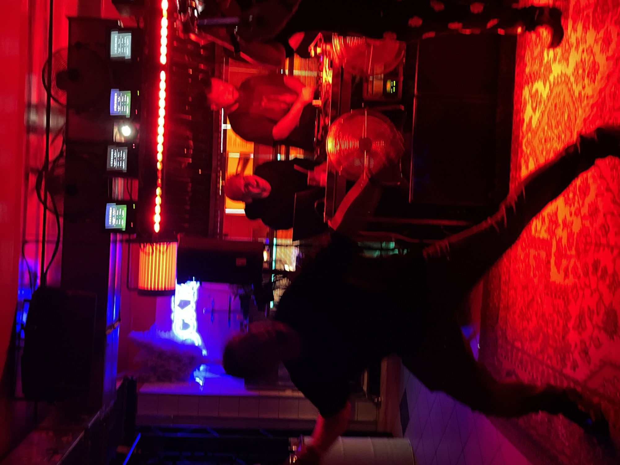 The image depicts an indoor scene within a bar or club, specifically identified as Jackie O Bar in Moscow, Russia, based on the visible blue neon sign displaying "JACKIE O" in the upper left background.
The environment is characterized by strong, pervasive red and blue artificial lighting. Red light dominates the foreground and midground, creating a heavily saturated atmosphere, while blue light accents the background and the left side.
In the foreground, a male individual, largely silhouetted, is captured in a dynamic pose, mid-movement, possibly dancing or gesturing. He wears a dark t-shirt and dark trousers, with his right leg bent and left leg extended. The floor beneath him is covered by a patterned rug, which appears red due to the lighting.
In the midground, two male individuals are positioned behind a DJ setup. The DJ on the right is actively operating turntables and a mixer, his hands on the equipment. The DJ on the left is partially obscured, observed near a laptop. The DJ booth features two large, circular, reflective objects, possibly cymbals or decorative elements, positioned vertically on either side of the main equipment. Below the DJ equipment, an audio component with green illuminated indicators is visible.
The background above the DJ booth features a row of five small monitors, each displaying text (consistent with "STREAMLABOR" visible in closer inspection) and having green status lights. Beneath these monitors, a horizontal strip of red LED lights spans the width of the area. To the right of the "JACKIE O" neon sign, a window with horizontal red blinds is visible. On the far right, a portion of another individual, wearing dark trousers with white polka dots, is visible. The ceiling area reveals mounted speakers and industrial fans. The scene captures a live DJ performance in a club setting.