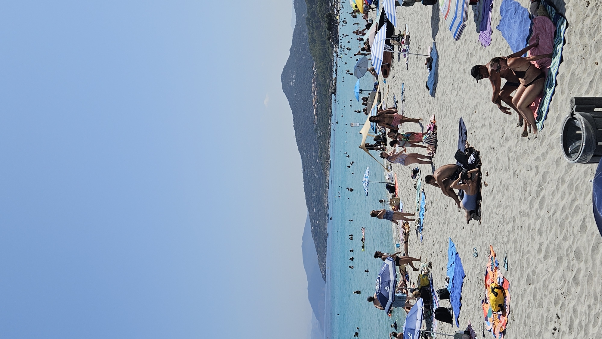 The image depicts a crowded beach scene in L'Île-Rousse, France, on a clear, sunny day. The turquoise water of the Mediterranean Sea stretches towards a hazy, tree-covered coastline and mountains in the background. Numerous people are scattered across the sandy beach, engaged in various activities. Some are sunbathing on towels and blankets, while others are swimming or wading in the shallow waters. Several beach umbrellas and a pop-up tent provide shade for beachgoers. Children are visible playing on the sand and in the water. Boats are dotted on the horizon. In the foreground, a trash bin is positioned on the sand. The overall atmosphere is one of a bustling summer beach day.