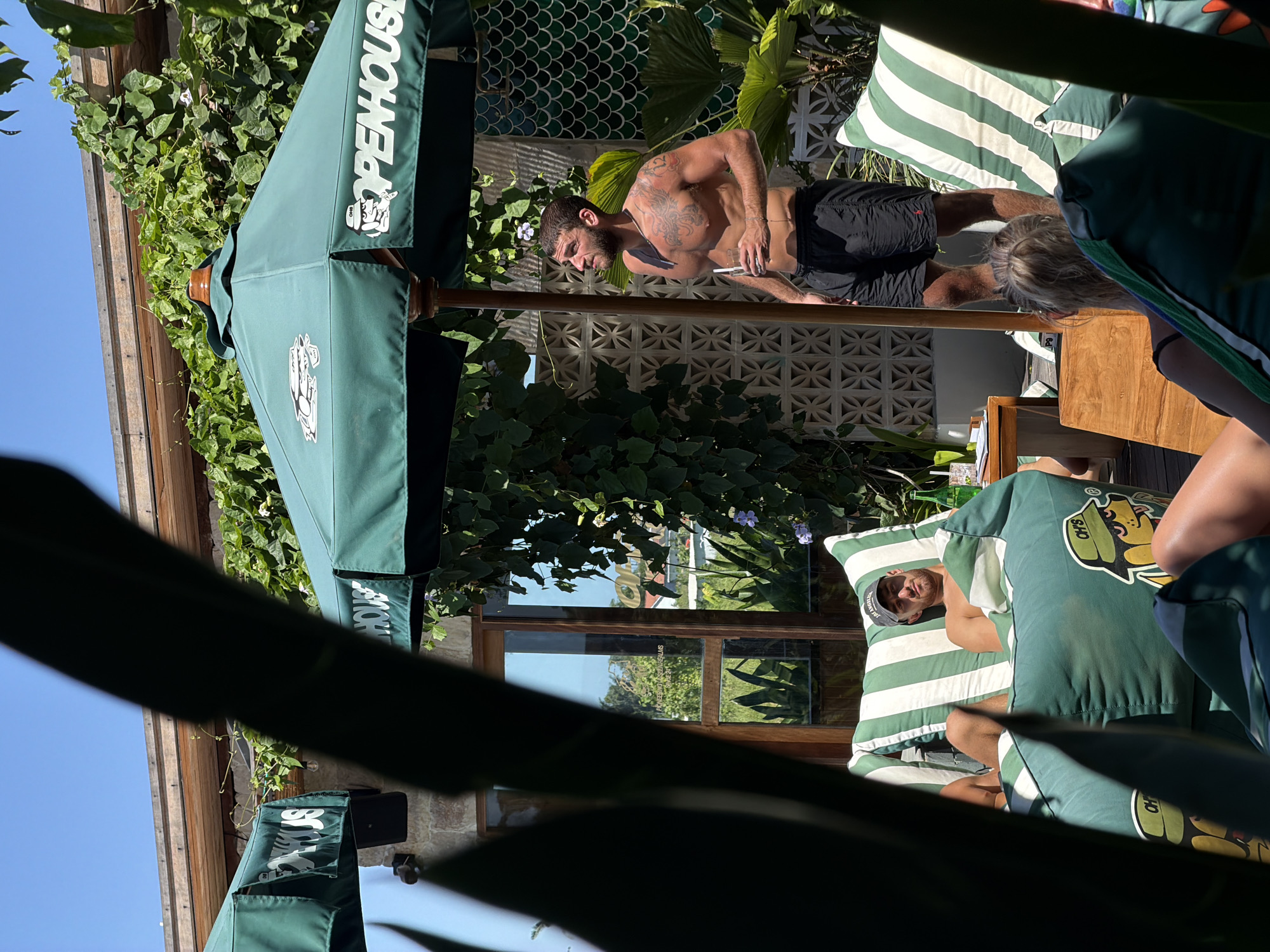 The image shows an outdoor lounge area, possibly in Mengwi, Indonesia, given the presence of tropical vegetation. A man with tattoos is standing and appears to be talking to someone whose back is turned toward the viewer. Another person is lying down on striped lounge cushions with pillows, appearing to be relaxing. There is a wooden table and outdoor furniture. A large green umbrella with the word "OPENHOUSE" printed on it provides shade. The setting seems to be a patio or deck area, with a decorative wall or fence in the background. Sunlight streams through the leaves and structures, creating strong shadows.