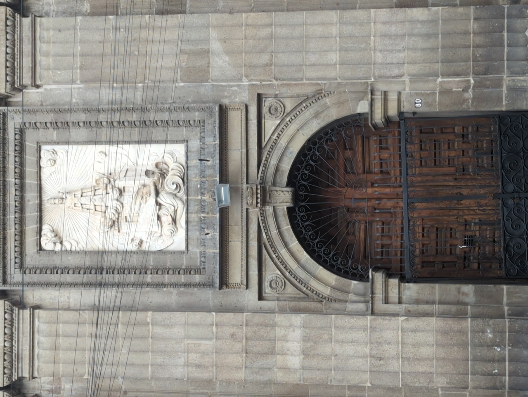 The image shows a close-up, overhead view of a building facade, likely in Mexico City, Mexico. The facade is constructed from grey stone blocks. Prominent features include a rectangular carved stone relief depicting religious figures, an arched doorway covered by decorative wrought iron grilles and a wooden door, and the number "80" on a stone block near the doorway. Multiple thin wires extend vertically across the facade.