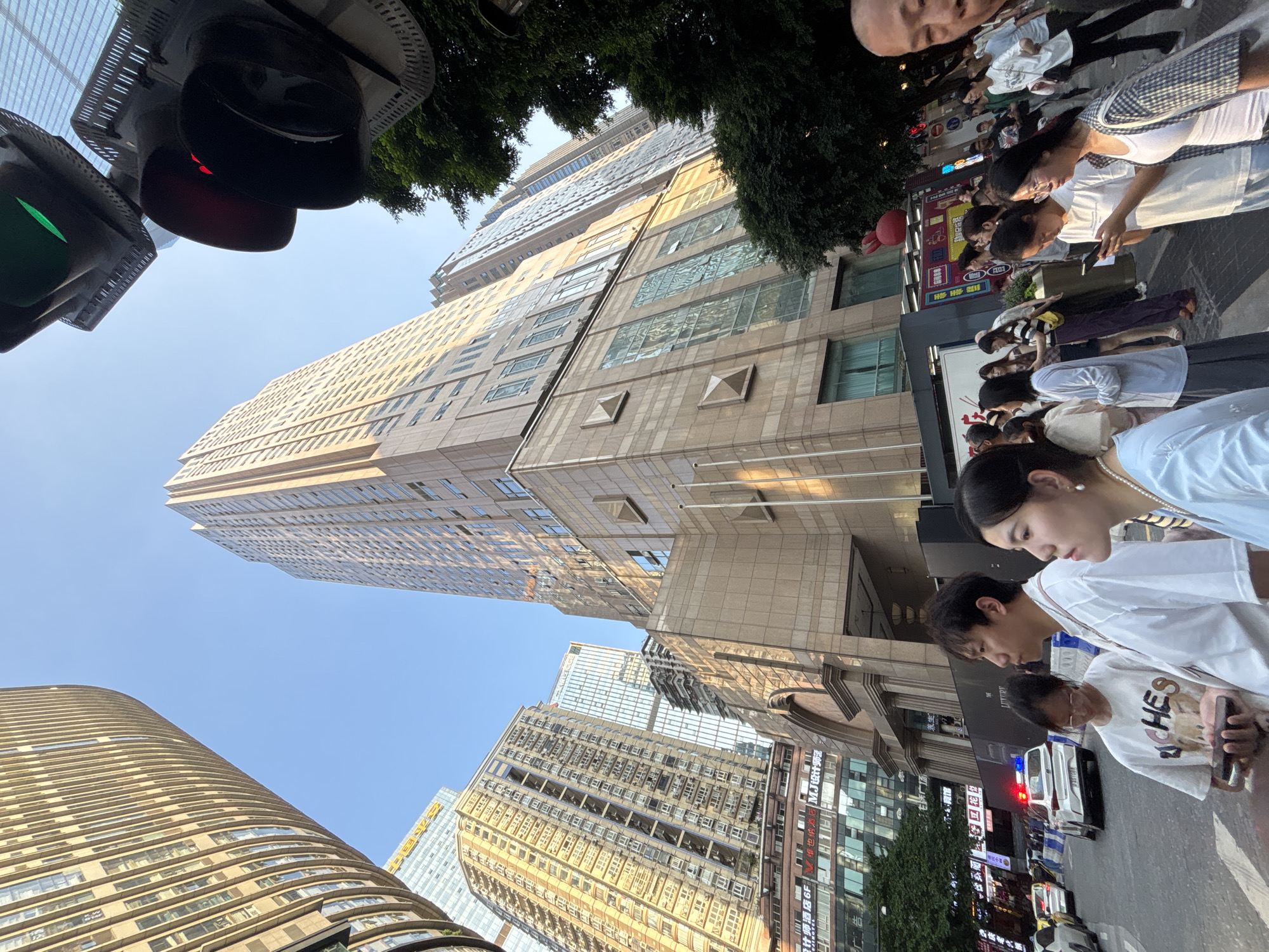 In an urban scene set in Yuzhong District, China, a diverse group of people are captured in motion, some standing and others walking along a sidewalk adjacent to a busy street. Towering modern skyscrapers, characterized by their glass facades and intricate architectural details, dominate the background, suggesting a bustling city environment. Sunlight bathes the scene, casting distinct shadows and highlighting the textures of the buildings, indicating it is likely daytime.
A traffic light, displaying a red signal, is visible, implying a momentary pause in the flow of vehicles. The people are dressed in casual, contemporary clothing, with some individuals looking at their phones or engaged in conversation. Their expressions range from neutral to mildly interested, conveying a sense of everyday life unfolding in a metropolitan area.
Several signs with Chinese characters are present on the buildings and storefronts, offering glimpses into the local commercial landscape. One sign displays "M.J.V Hotel," indicating a lodging establishment. Another sign with yellow text reads "万客隆酒店" (Wan Kelong Jiudian), which translates to "Wan Kelong Hotel." The overall atmosphere is one of a vibrant urban center during daylight hours.