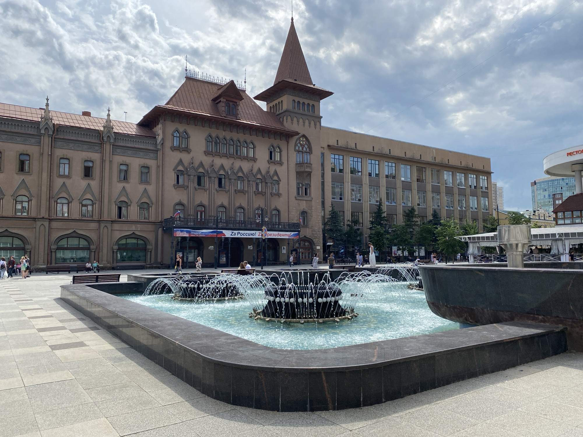 In the city of Unknown, Unknown, a large, multi-tiered fountain with a dark stone border dominates the foreground of the image. The fountain's water jets are actively spraying, creating a frothy, luminous surface. Behind the fountain, a wide, paved plaza extends towards an imposing building. This building, with its ornate facade, reddish-brown tiled roof, and a prominent tower with a conical top, displays architectural elements suggesting a historical or governmental purpose. Several arched windows and decorative stonework are visible on its exterior. To the right of this main building, a more modern, multi-story structure with large, reflective windows can be seen. The sky above is partly cloudy, with dramatic, textured clouds casting shadows. Several people are scattered across the plaza and in front of the buildings, some walking, others seated on benches or observing the fountain. Banners with Cyrillic text are displayed on the facade of the historical building. Trees line the area in front of the modern building, and a circular structure with signage that reads "RECTO" is visible in the distance on the right.