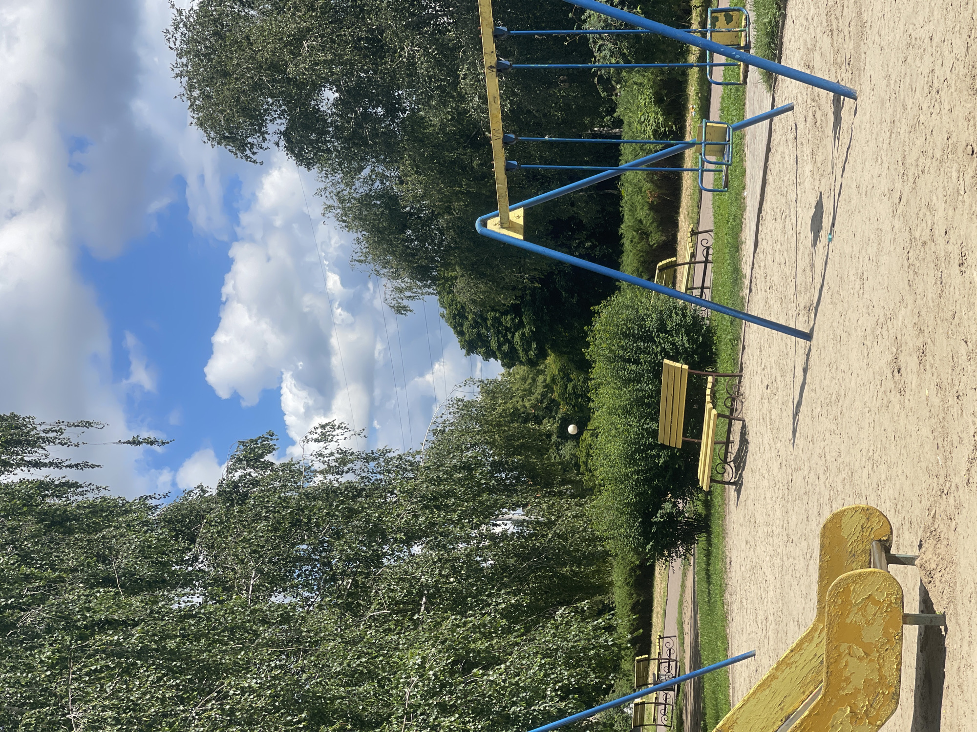 An outdoor playground is depicted on a sunny day with a blue sky filled with scattered white clouds. The playground features a sandy ground with several swings, a slide, and park benches. In the foreground, a worn yellow slide is visible, and further back, a set of swings with blue metal frames and yellow seats are present. Two yellow park benches are also situated in the playground area, one of which is near a dense green bush. Tall trees with green foliage surround the playground, creating a natural setting. Power lines are faintly visible in the background, suggesting the playground is located within a populated area. The overall scene is peaceful and recreational.
