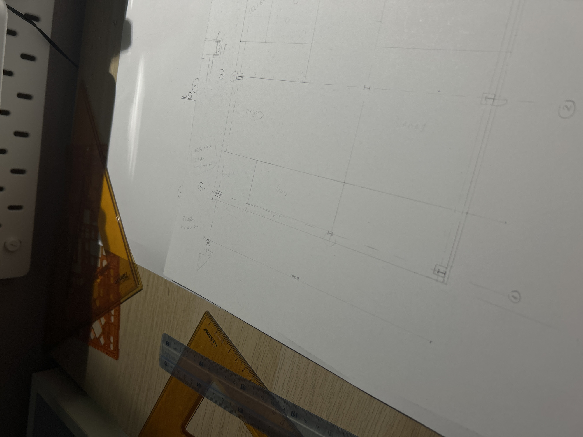 This image shows a drawing on a white piece of paper laid on a wooden surface. The drawing appears to be a plan or diagram, possibly architectural in nature, with lines, shapes, and handwritten annotations in what seems to be Bulgarian. An "Aristo" brand set square and ruler are positioned on the paper, presumably used for drawing and measuring.
There is no information provided to connect the image to Sofia, Bulgaria, or to identify any people or subjects. The only identifiable objects are the drawing, paper, ruler, set square, and the wooden surface.