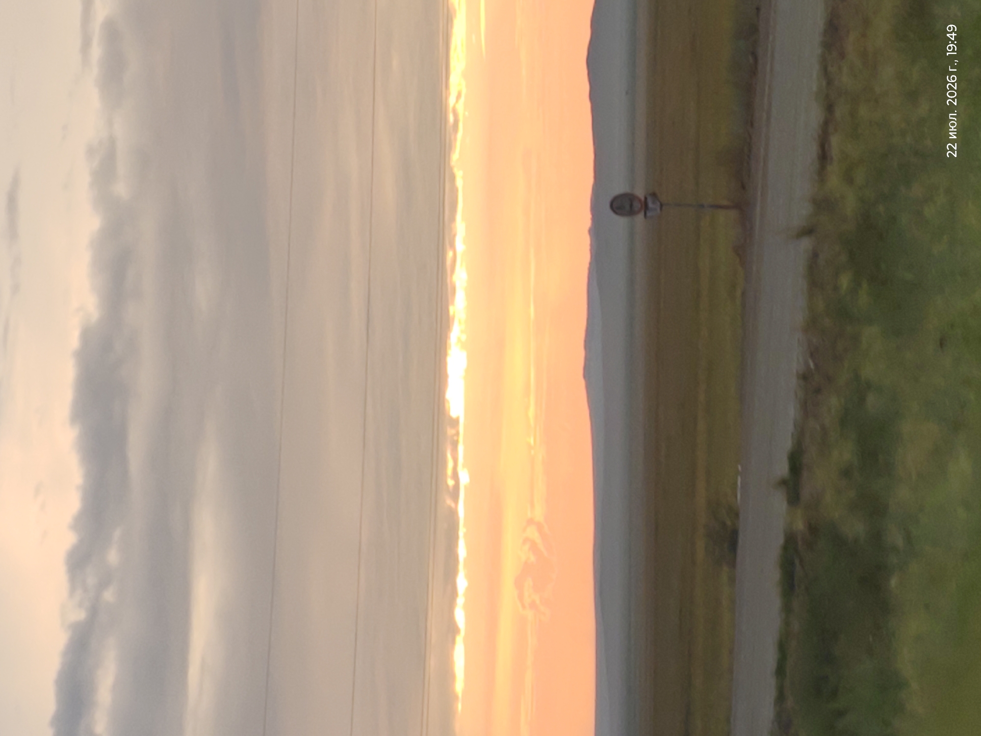 A landscape photo captures a sunset over a flat, grassy plain. The sky is filled with hues of orange and yellow, with a band of bright light near the horizon where the sun is setting. Wispy clouds drift across the sky, and power lines are visible in the upper portion of the frame. In the middle ground, a dirt road curves through the landscape, with a single traffic sign standing beside it. The foreground is dominated by green grass and vegetation. The timestamp "22 июл. 2026 г., 19:49" indicates the date and time the photo was taken.