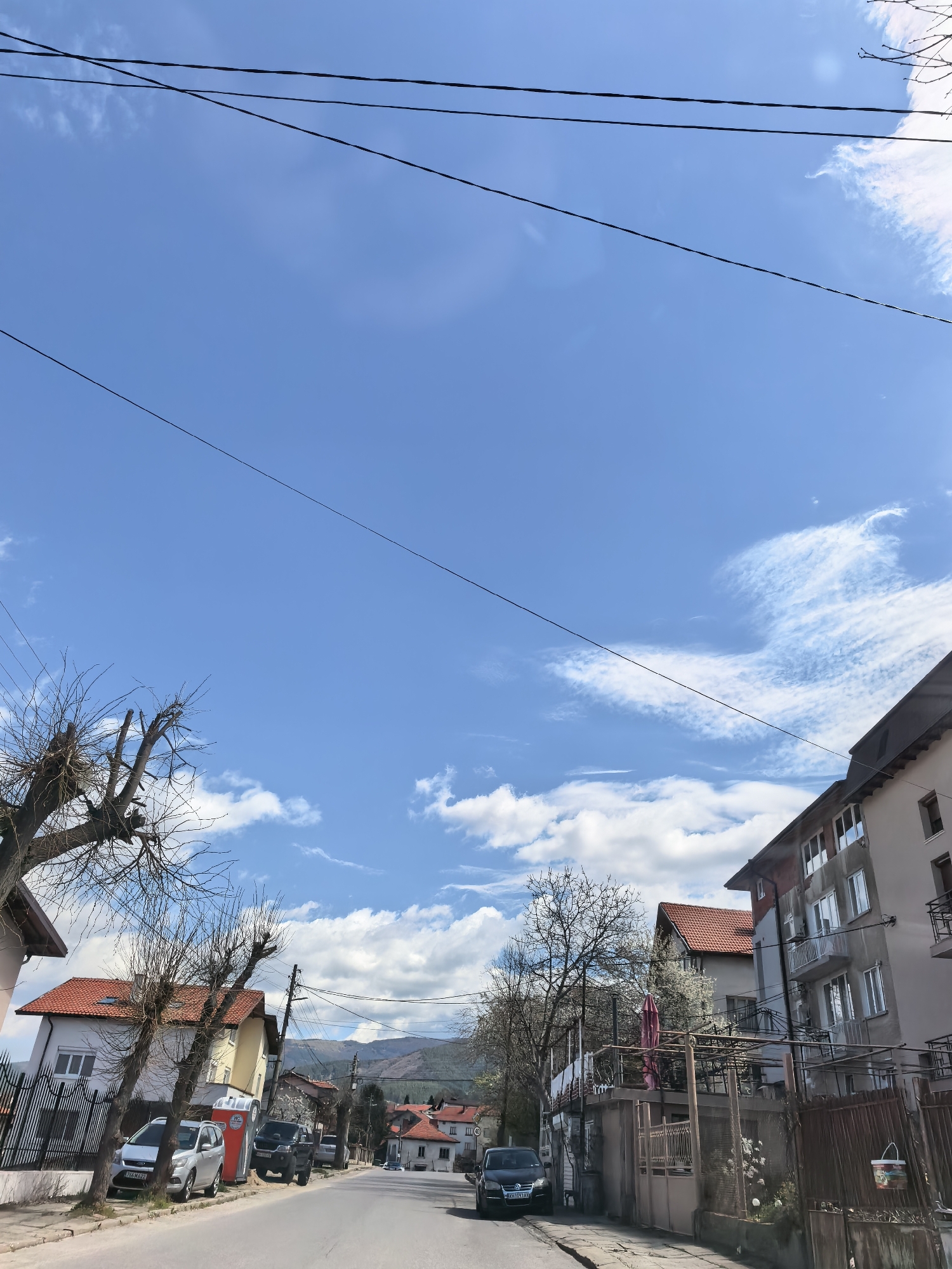 The image shows a street in an urban area. On either side of the road are buildings with red tile roofs. A car is parked on the left side of the street, and another car is parked on the right side of the street. There are several trees on either side of the road, and some of them are bare. In the background, there are hills and mountains. Overhead, there are several power lines stretching across the sky. The sky is blue with some clouds. The location is Unknown, Unknown.