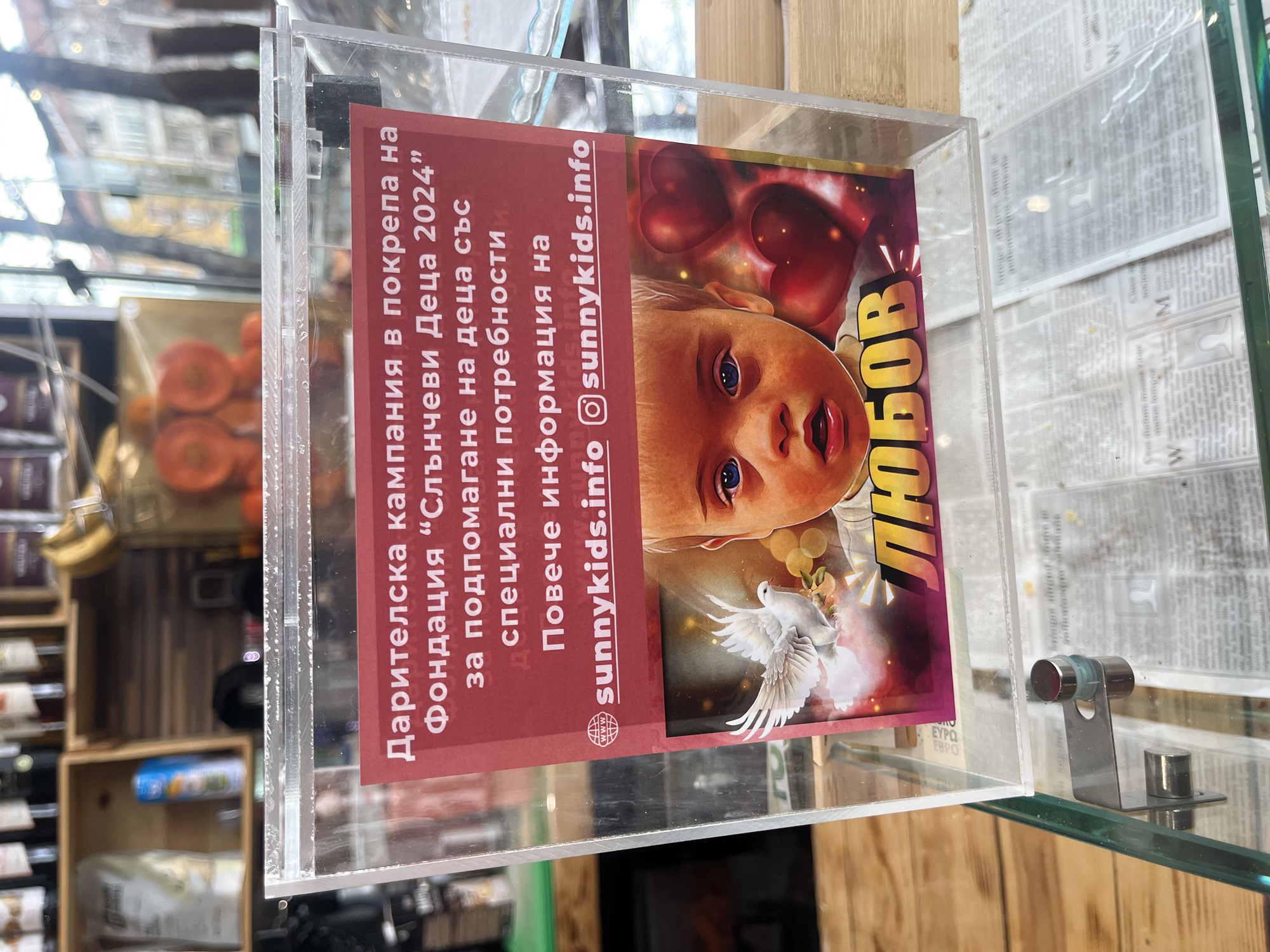 The image shows a transparent donation box with a promotional poster inside. The poster advertises a fundraising campaign for the "Sunny Kids 2024" foundation, which supports children with special needs. The poster provides information on the website sunnykids.info and the Instagram account @sunnykids.info. The poster also features an image of a baby with hearts in the background, as well as a dove. The box is placed on a glass surface, and a newspaper is underneath. In the background, shelves are visible with various items for sale. This scene may be in a retail location, such as a store or café in Sofia, Bulgaria.