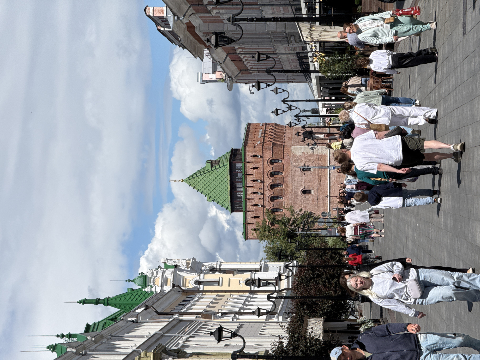 The image captures a sunny day on Bolshaya Pokrovskaya Street in Nizhny Novgorod, Russia, a bustling pedestrian thoroughfare. A prominent brick tower with a green roof, likely part of the Nizhny Novgorod Kremlin, dominates the background, serving as a historical landmark.
The street is lined with elegant buildings, featuring ornate architecture with green accents on their roofs. The foreground is populated by a diverse group of people walking in both directions. Some individuals are captured mid-stride, engrossed in conversation or carrying shopping bags. Others are observing the surroundings or posing for photographs.
The scene is characterized by a lively atmosphere, with people enjoying the urban landscape and the warm weather. The presence of street lamps and trees suggests a well-maintained public space designed for leisure and recreation. The overall impression is one of a vibrant city center, blending historical charm with contemporary urban life.