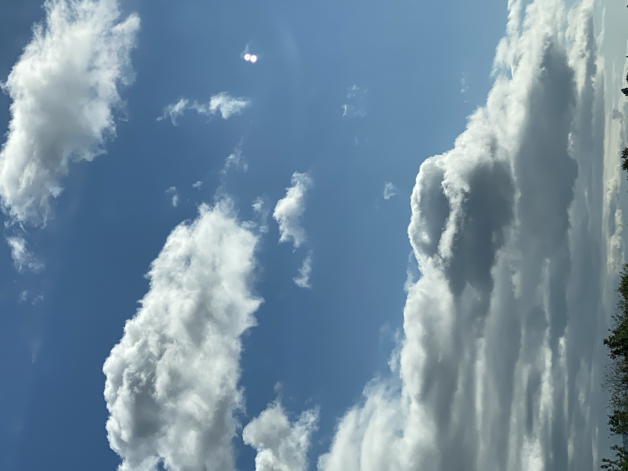 The image shows a bright blue sky filled with scattered white and gray clouds. In the upper right quadrant, two bright, circular lens flares are visible. A thin line of green foliage is visible at the very bottom of the image, suggesting a terrestrial landscape beneath the sky. The scene is outdoors. There are no people or specific structures of Heringen/Helme, Germany identifiable in the image.