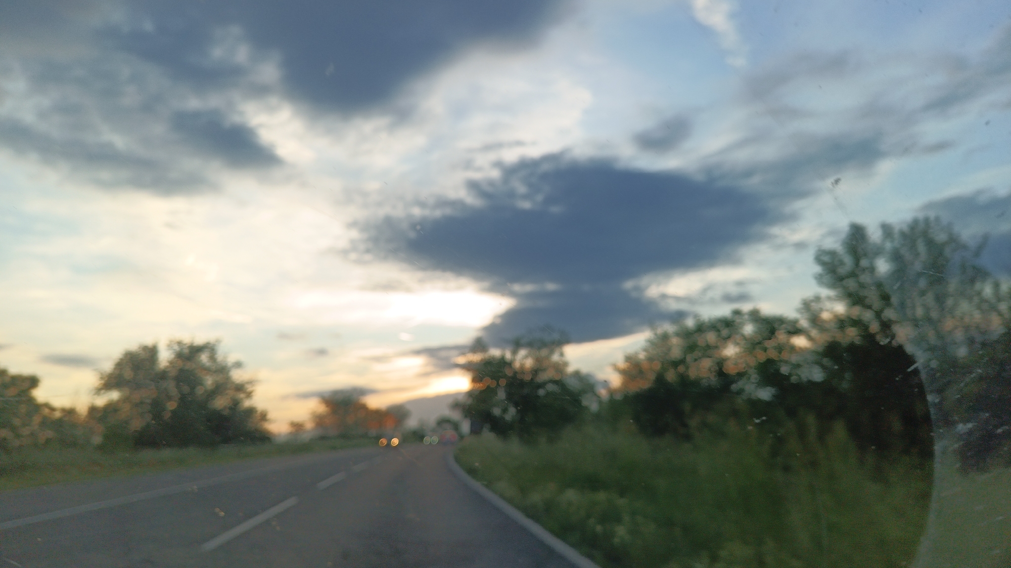 The image shows a blurred view from inside a vehicle driving on a road, likely during sunset or twilight. The road has two lanes and a white line marking the center. The surroundings include roadside vegetation and scattered trees. The sky contains a mix of blue and gray clouds with a hint of sunset colors. There are a few distant car lights visible on the road ahead. The windshield appears to be dirty with some smudges or scratches. It is not possible to confirm the location as Burgas, Bulgaria, or any specific city without further information.