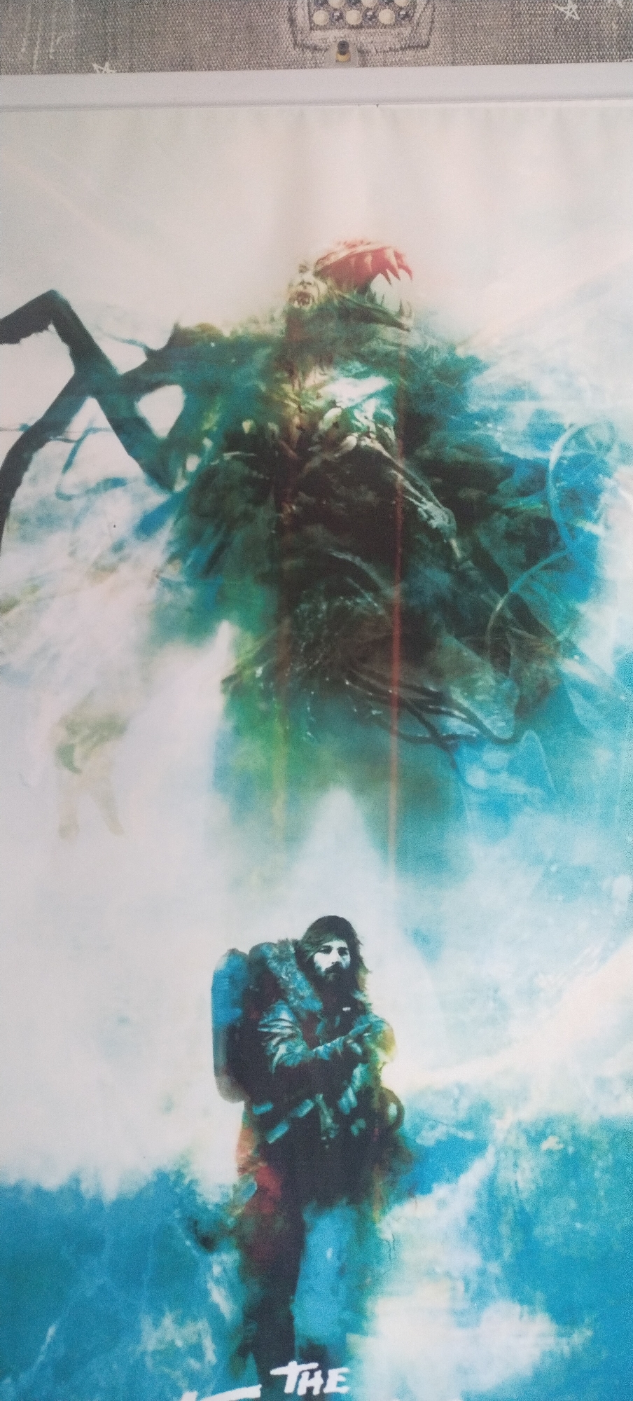 The image depicts a poster or artwork featuring a fantastical scene. In the foreground, a man with a beard, wearing a heavy, fur-lined jacket and a backpack, is positioned in a kneeling or crouched stance. He appears to be looking upwards. Behind him and above, a large, monstrous creature is depicted, rendered in dark, swirling greens and blues with hints of red and white. The creature has sharp teeth and appears to be made of organic, possibly alien, material, with tendril-like appendages. The background is a hazy, atmospheric mix of white and blue, suggesting a cold or ethereal environment. The bottom portion of the image shows text, with "THE" clearly visible, suggesting it is part of a title. There is no discernible connection to the city of Pisochyn, Ukraine, as the image is purely illustrative and fantastical.
