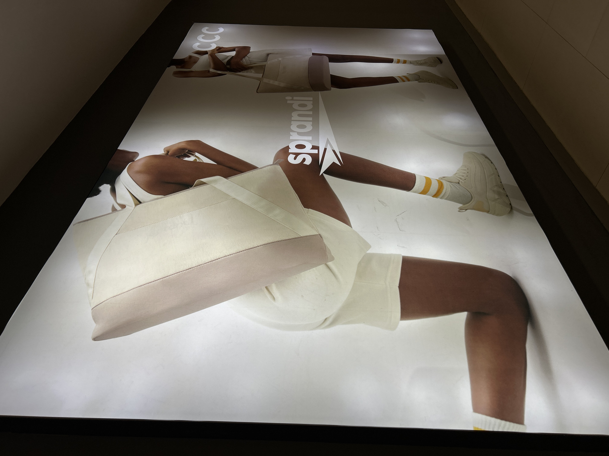 The image shows a brightly lit advertising panel, likely located in Plovdiv, Bulgaria, featuring a promotional advertisement for the brand “sprandi” and "CCC". The advertisement shows a person lying down, facing the viewer, dressed in a white outfit, including what appears to be a tank top, shorts, and socks with yellow stripes, with a pair of white sneakers. They have a light-colored tote bag next to them. Above this image, there is a smaller version of the same model, seemingly mirrored or repeated, which adds a dynamic element to the advertisement. The overall scene suggests a fashion-related or retail environment.
