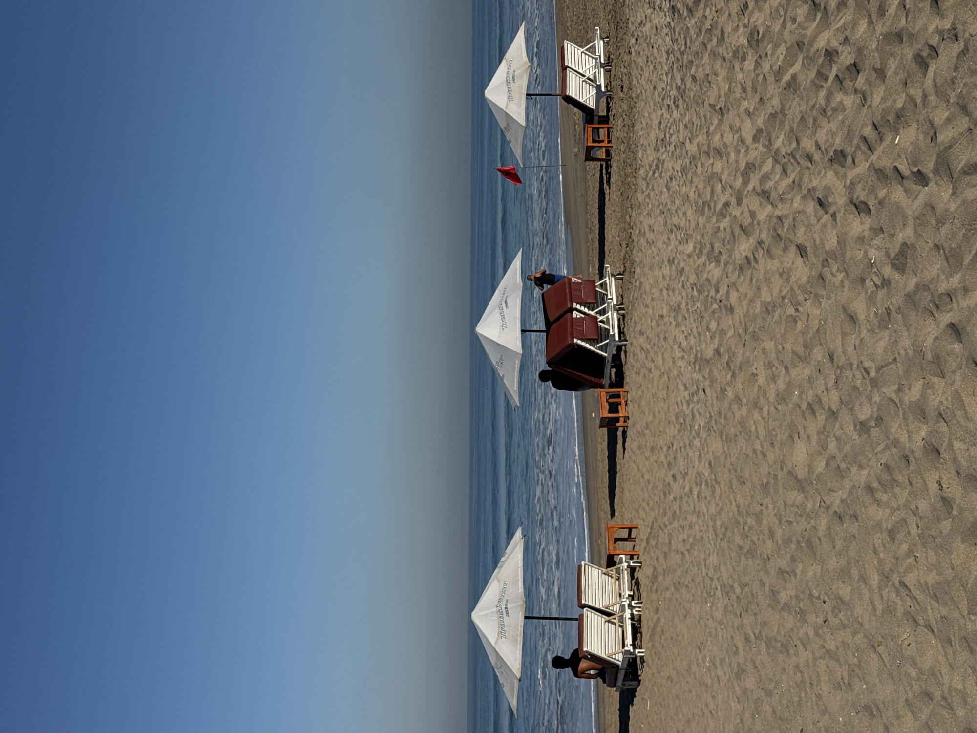 Here's a description of the image:
The photograph presents a beach scene featuring a sandy shore adjacent to a body of water, presumably the ocean. The sky is clear blue.
The beach contains rows of beach umbrellas and sun loungers, some of which are occupied by people. The sand appears to be dark in color.
The scene appears to be taken during daytime under clear skies. The location is not evident from the image itself.