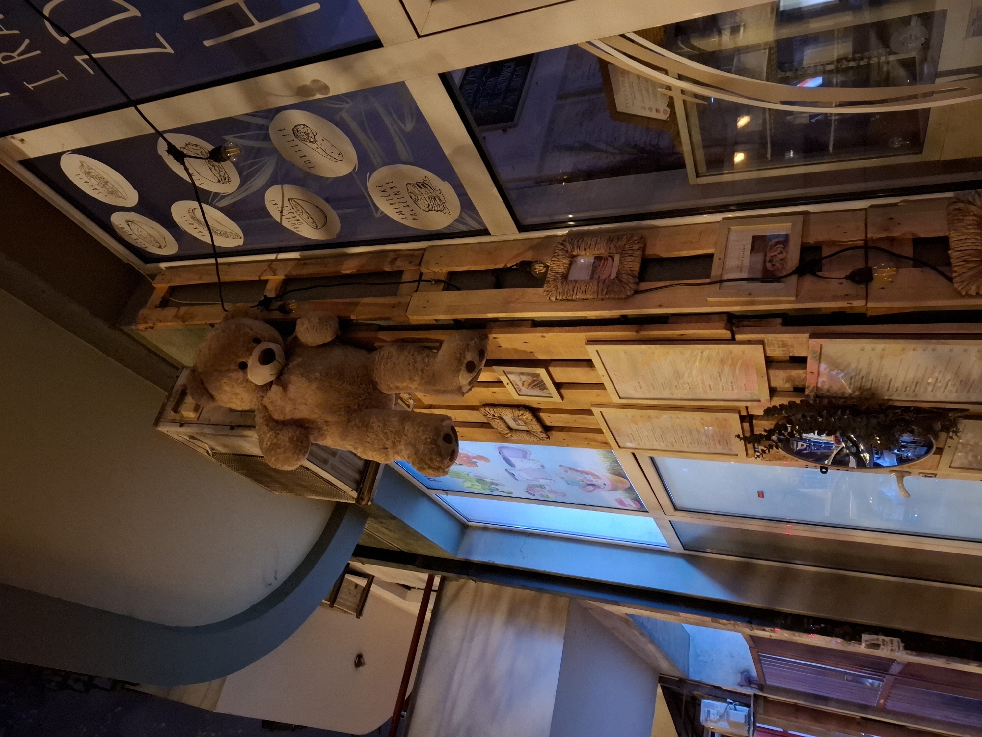 A teddy bear hangs from the ceiling of a restaurant in Novi Sad, Serbia. The ceiling is decorated with wooden pallets, framed pictures, and menus. Some windows are decorated with pictures of food. The restaurant interior is modern, with curved walls.