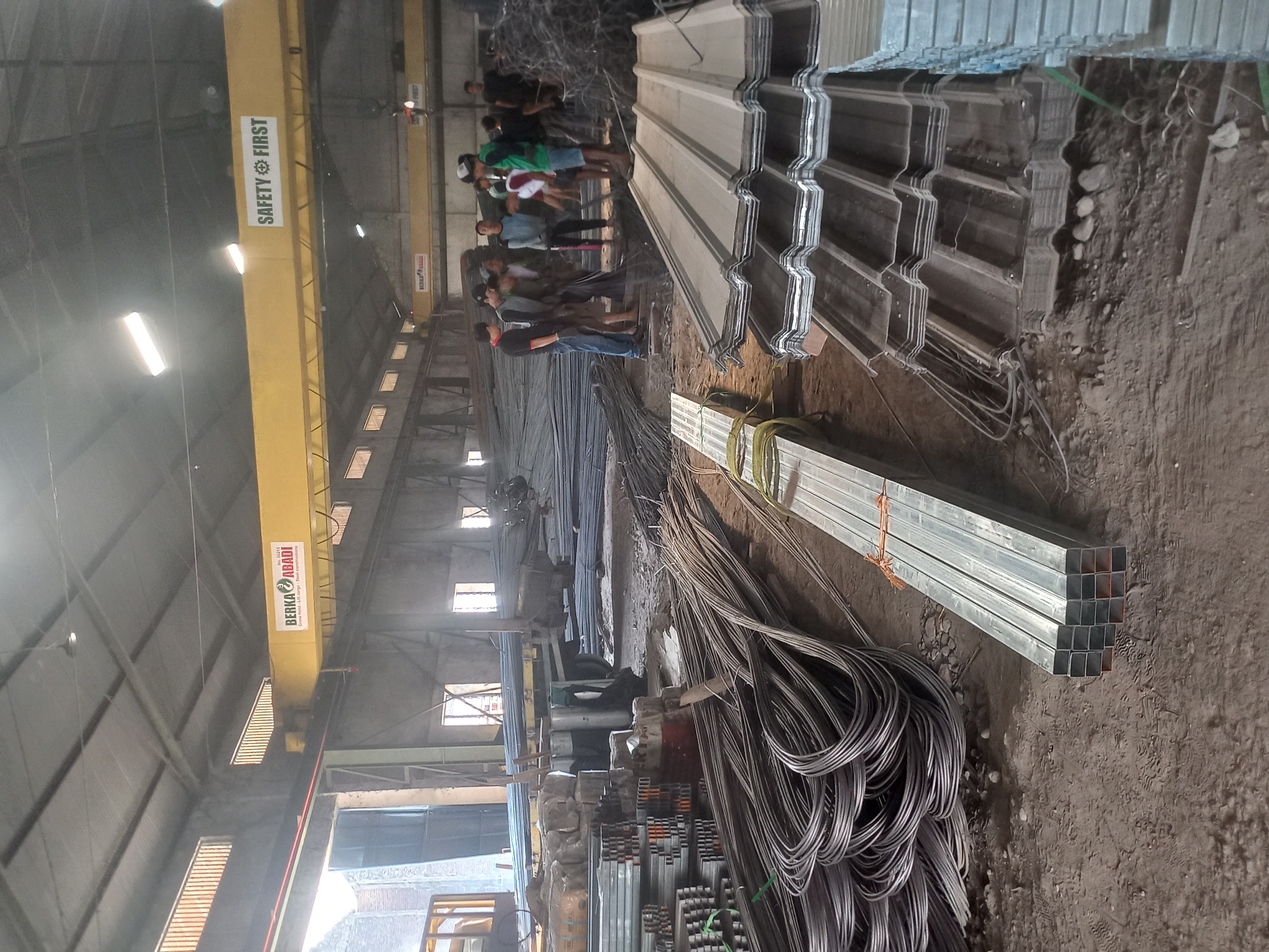 The image depicts the interior of a warehouse in Kacangan, Indonesia, likely a steel or construction supply facility. A large yellow overhead crane with signage "BERKAB ABADI" and "SAFETY FIRST" is visible. The warehouse floor is dirt and concrete, cluttered with various construction materials. In the foreground, stacks of rebar are coiled and uncoiled, alongside bundles of square metal tubing and corrugated metal roofing sheets. In the middle ground, several men, dressed casually in t-shirts and shorts, are gathered around a truck laden with steel bars, appearing to be workers or customers. The environment suggests an active industrial setting with abundant building materials.
